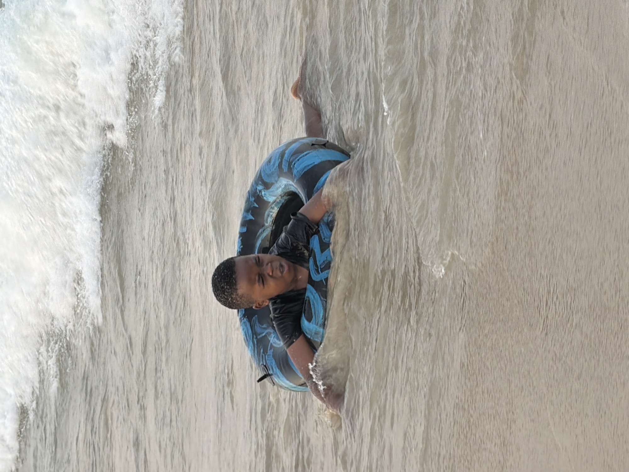 An African child, wearing a black t-shirt, is in the ocean at an unknown beach in Dar es Salaam, Tanzania, using a blue and black inflatable ring for flotation. The child's face is contorted in an expression of discomfort, with their mouth open. Waves are breaking behind the child, creating white foam. The water is a light brown color, indicative of sand stirred up from the seabed. The child is holding onto the sides of the inflatable ring, and their legs are submerged beneath the water, with one leg visible extending to the right. The scene depicts a moment of child playing in the sea.