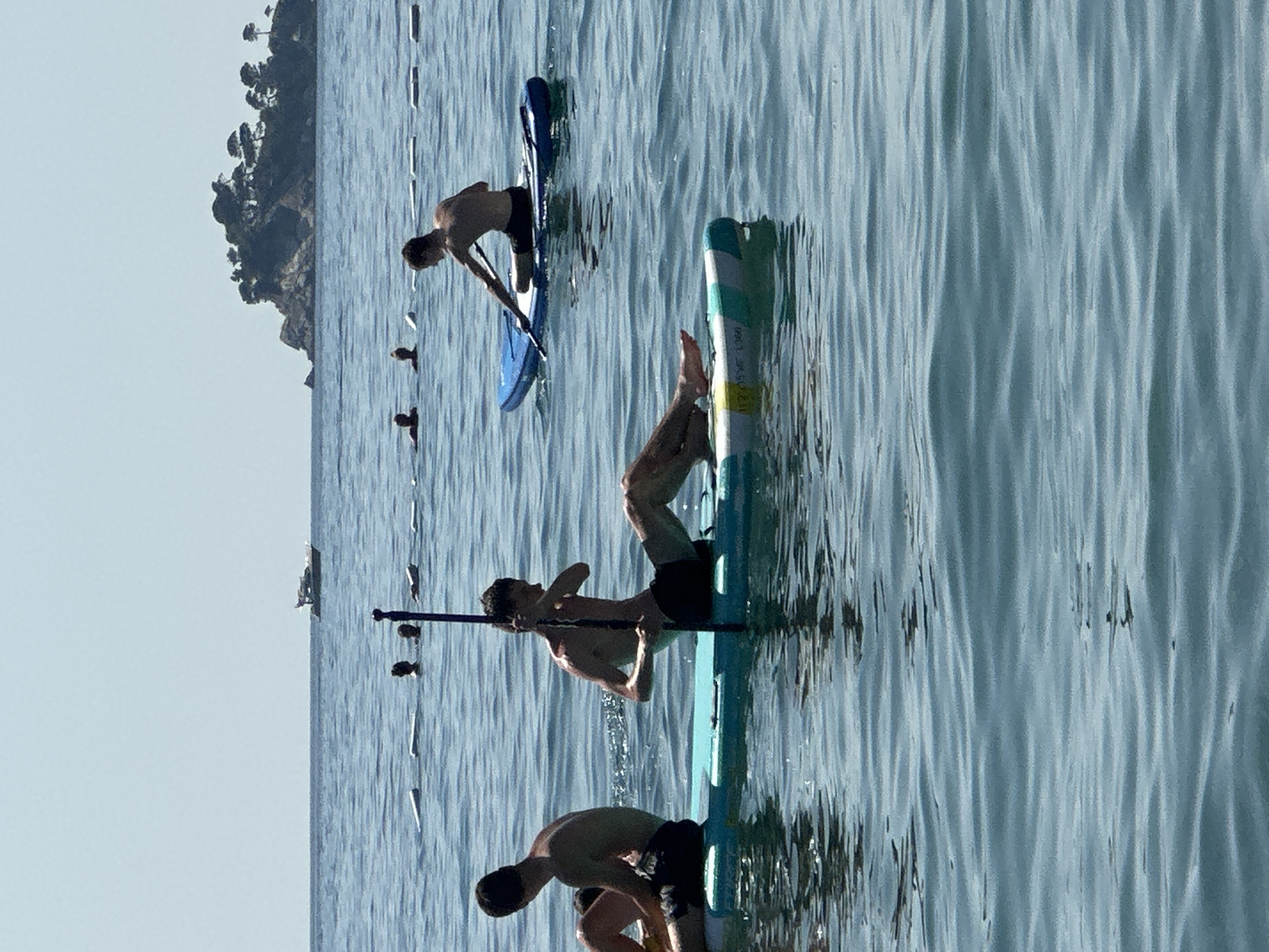 In the city of Đurovići, Montenegro, on a bright, sunny day, several individuals are enjoying water activities. In the foreground, two young men are seated on separate paddleboards. The man on the left, wearing dark shorts, is facing away from the camera. The man on the right, also in dark shorts, is holding a paddle and facing left. Behind them, several other individuals are visible in the water, with only their heads above the surface. In the background, a small boat is seen on the horizon, and a rocky island covered in trees is to the right. The sea is calm with gentle ripples, and the sky is clear.