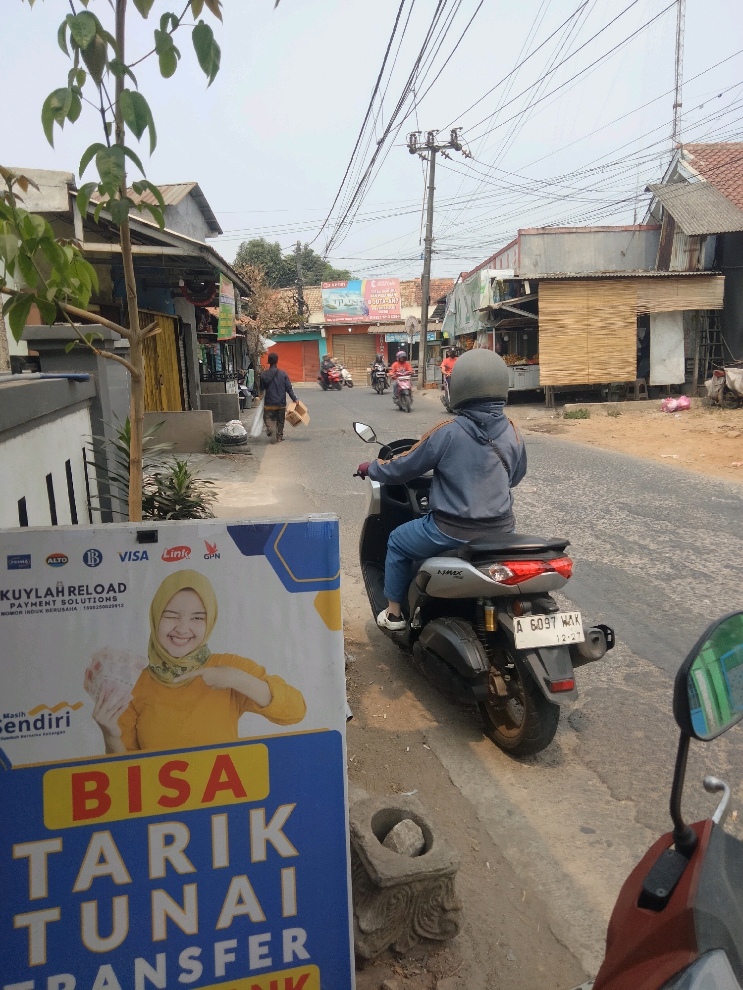 The image depicts a street scene in Suka Sari, Indonesia. In the foreground, a person wearing a gray helmet and a blue hooded jacket is riding a dark-colored motorcycle. The motorcycle has a license plate reading "A 6097 WAK 12-27". To the left, a white sign with blue and yellow accents features an advertisement for "KUYLAR RELOAD PAYMENT SOLUTIONS" and shows an image of a smiling woman in a yellow hijab holding money. The background shows a street lined with buildings, some with corrugated metal roofs and bamboo accents. Power lines crisscross above the street. In the distance, other motorcycles and pedestrians are visible, suggesting a moderately busy environment. The overall atmosphere appears to be a typical day in a residential or commercial area.