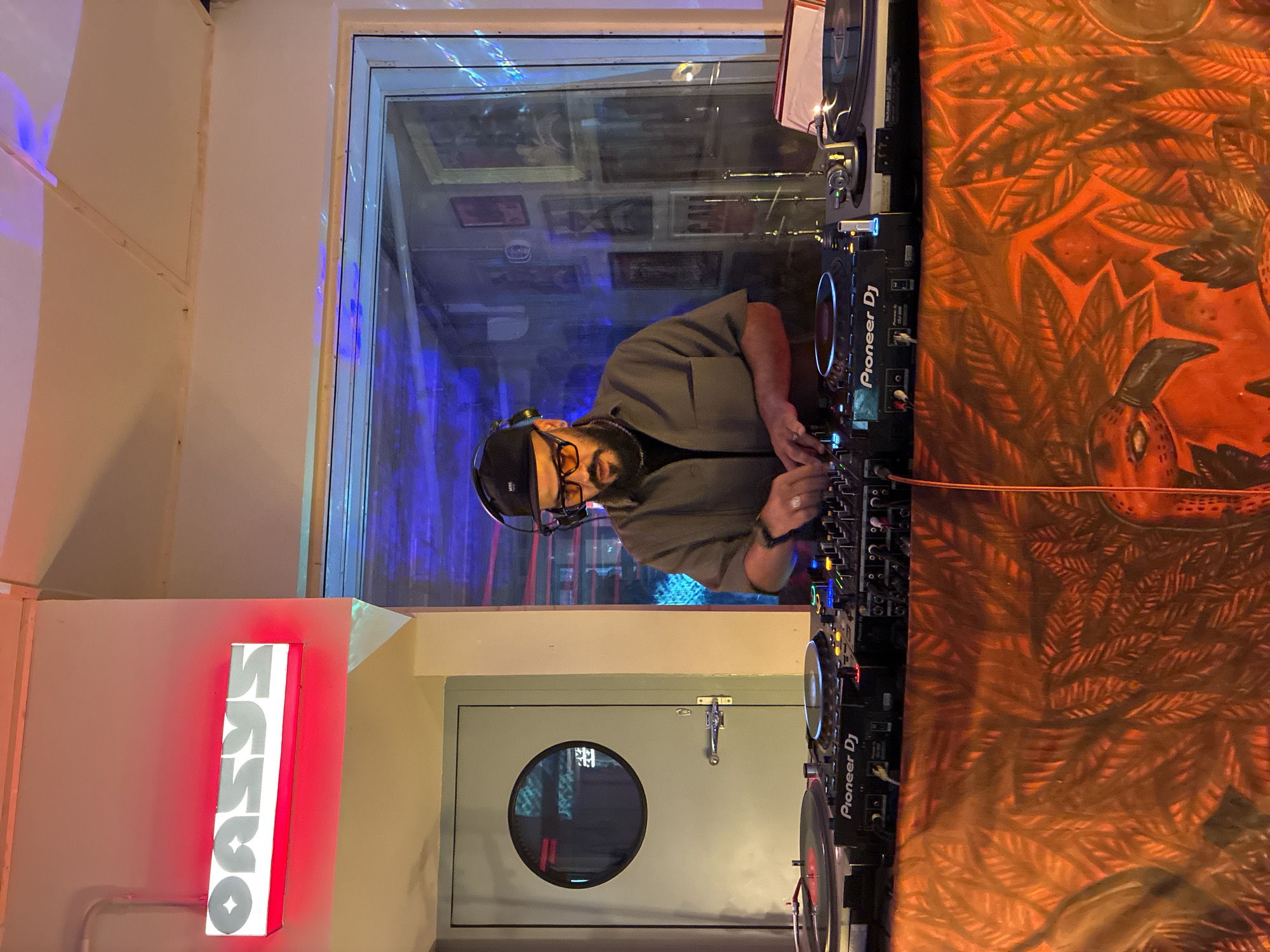 An indoor setting reveals a DJ booth where a man with a beard and glasses, wearing a cap and headphones, is actively DJing using a Pioneer DJ console. To his right, a mural displays orange hues and features bird-like patterns. To his left, a bright red neon sign is visible, displaying text in a mirrored fashion. Behind him, a window reflects interior elements including framed art and other lighting. A door with a circular window is also in view. While the image's exact location in Barcelona is not apparent, the presence of Spanish text on the sign indicates a Spanish-speaking region.
