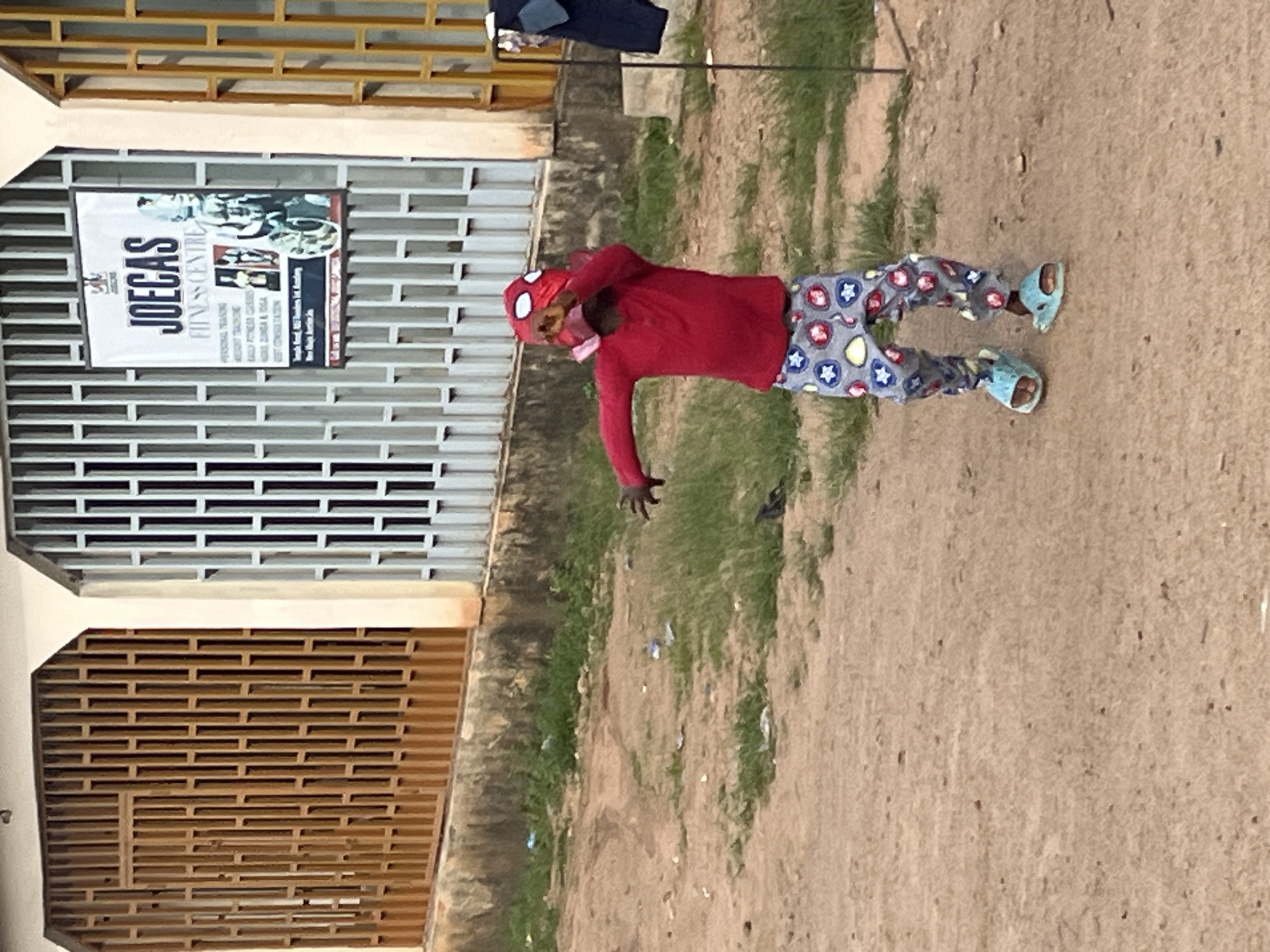 A child wearing a red long-sleeved shirt, patterned pants, and blue slide sandals stands in a dusty, unpaved area. The child has a red and white mask resembling Spider-Man's mask on their head, with the mask partially covering their face. The child's left arm is raised, and their right arm is extended to the side, as if in motion. Behind the child is a building with a light-colored facade and metal security grilles, one of which has a white sign that reads "JOECAS FITNESS CENTRE." To the right of the building, dark clothing is hanging on a clothesline or rack. The ground in front of the building is a mix of dirt and sparse green grass, with some scattered litter. The scene appears to be outdoors in a semi-urban or rural setting, possibly in Barakin Akawo, Nigeria, given the architectural style and environment.