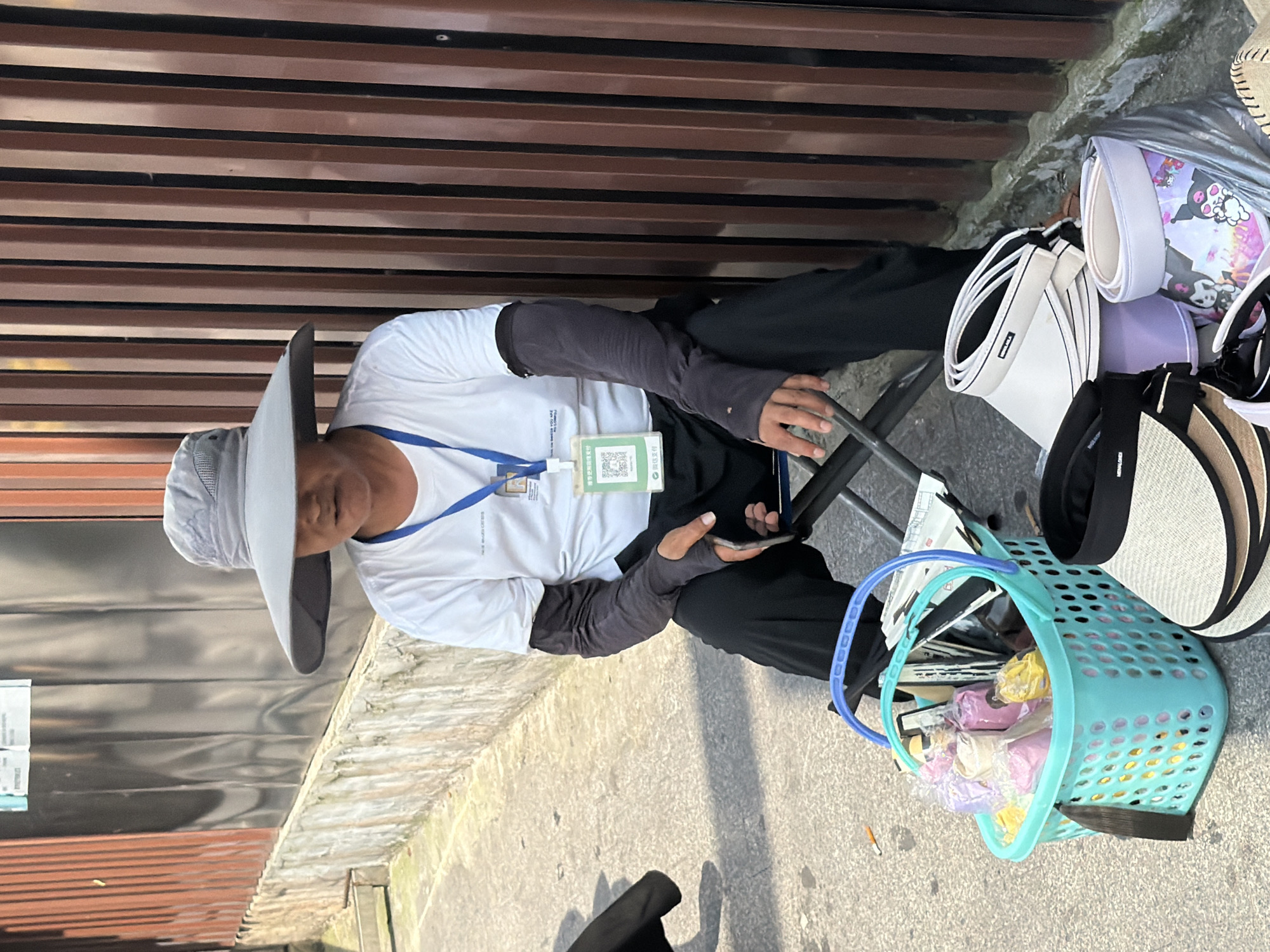 A person sits on a folding chair in front of a vertical slatted fence. They are wearing a wide-brimmed grey hat, a white t-shirt with long dark sleeves underneath, and black pants. A lanyard with a badge is visible around their neck. Next to them, on the concrete ground, is a bright turquoise plastic basket filled with various items wrapped in plastic, including what appear to be rolls of fabric or paper in pink and yellow. In front of the basket, several visors are stacked, some white and cream, and one with a cartoon character print. The overall setting suggests an outdoor market or street-side stall in Shapingba District, China, given the presence of merchandise and the urban environment.