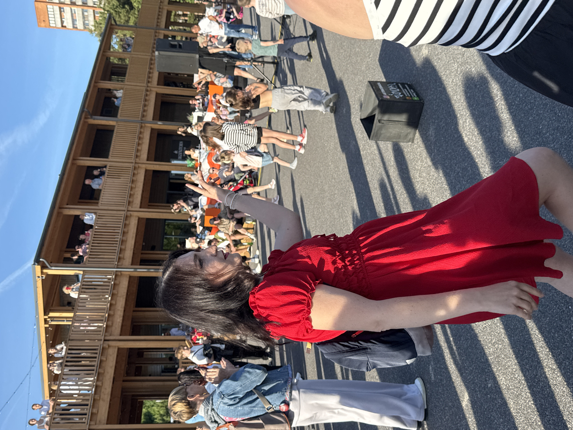 The image depicts an outdoor event likely taking place in Tallinn, Estonia, possibly at Telliskivi Creative City, an area known for its vibrant atmosphere and public gatherings. In the foreground, a woman in a bright red dress is dancing or gesturing with her right hand raised. Behind her, a person wearing a denim jacket and light-colored pants is also in motion, possibly clapping. The background reveals a multi-story wooden building with balconies occupied by spectators. Several children are visible on the paved ground, some dancing or observing. A black speaker stands on a tripod to the right, and a black cube-shaped bin is on the ground. The scene is illuminated by natural daylight under a clear blue sky with wispy clouds.