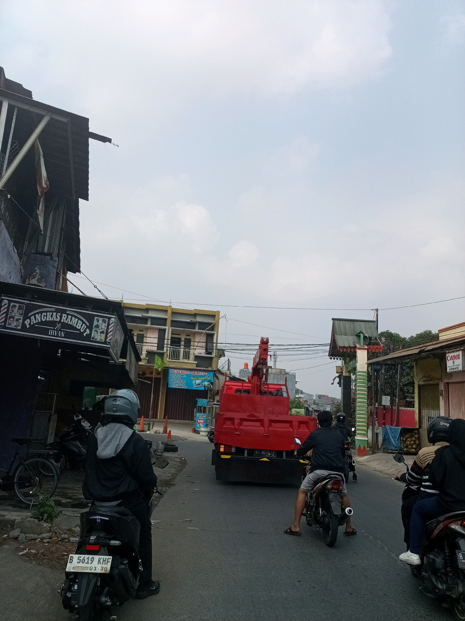 In Bekasi, Indonesia, a street scene unfolds with a bright, overcast sky. A red truck with an extended crane is the dominant vehicle, positioned on the right side of the road. Several motorcycles navigate the asphalt. In the foreground, a person wearing a grey helmet and black jacket rides a black motorcycle, license plate B 5619 KHF visible. Further back, another motorcyclist in a black jacket and grey shorts is also on the road, as is a third person on a motorcycle, wearing black. On the left side of the street, a building with a prominent sign reading "PANGKAS RAMBUT IRVAN" (barber shop) is visible. Adjacent to it, another building has a sign indicating "MENERIMA SERVIS TV LCD..." (service TV LCD). The environment is urban, with a mix of commercial and residential structures lining the street.