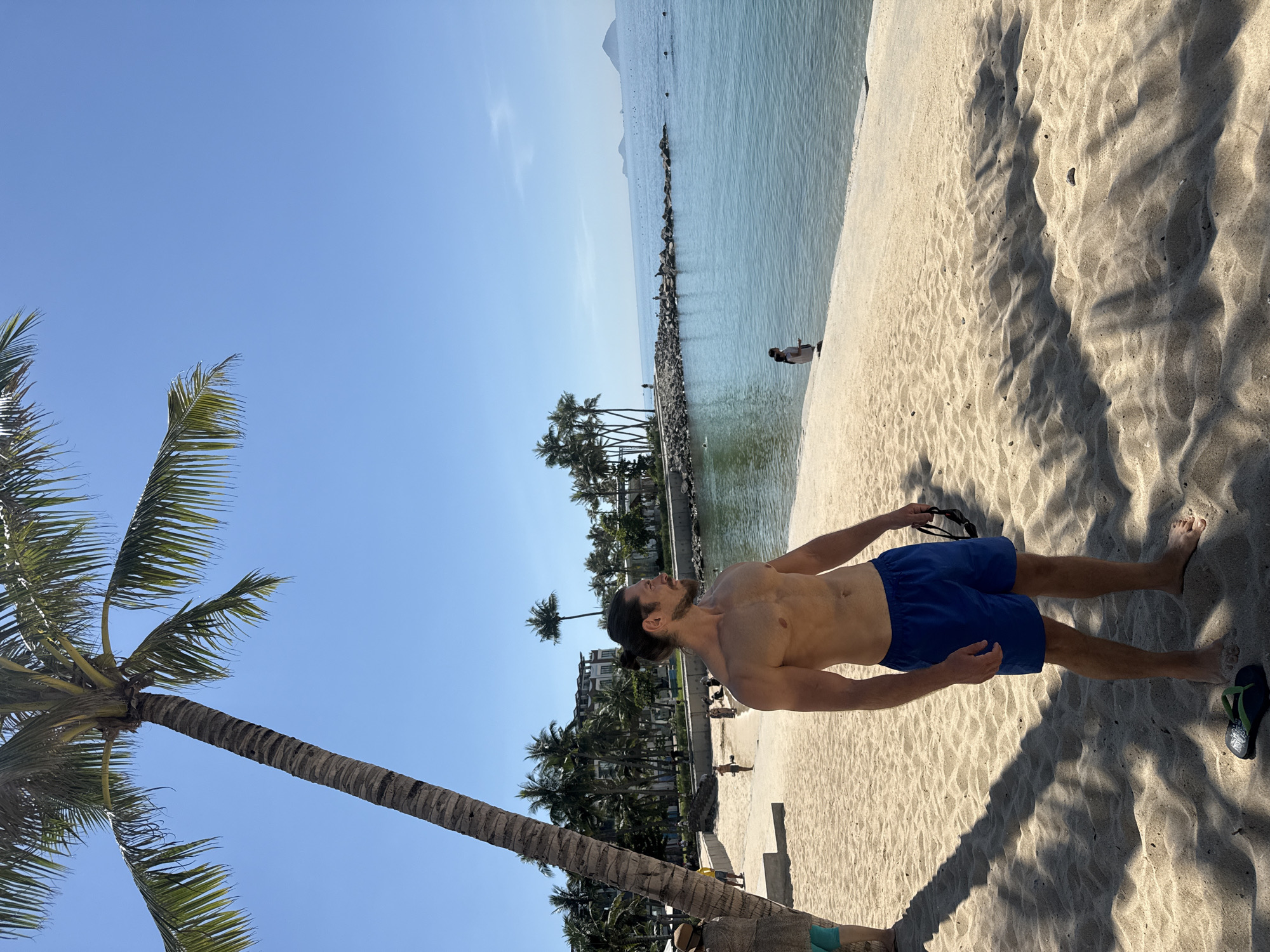 A man is standing on a sandy beach, looking out at the ocean. He is shirtless and wearing blue swim trunks. There is a palm tree to his left, and the ocean stretches out before him, with several small islands in the distance. In the background, there are some buildings and more palm trees. The sky is clear and blue. The man is holding a pair of sunglasses in his right hand. A pair of flip-flops is on the sand near his feet. The sand is light brown and appears to be a beach. The water is clear and a light blue color.