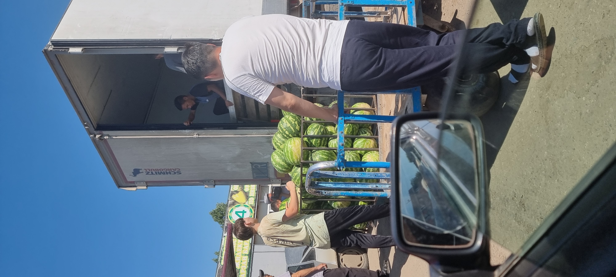 In Aktobe, Kazakhstan, under a clear blue sky, a large white cargo truck is parked. The rear of the truck is open, revealing a person inside. In front of the truck, a blue metal cart is filled with numerous watermelons. Several individuals are present around the cart and the truck. One man, wearing a white t-shirt and dark pants, stands with his back to the camera, looking towards the truck. Another younger man, in a light-colored t-shirt and dark pants, is positioned behind the watermelon cart. A third person, in a dark t-shirt, is visible within the open truck, seemingly engaged in handing items out. The scene appears to be a commercial setting, possibly a market or distribution point.