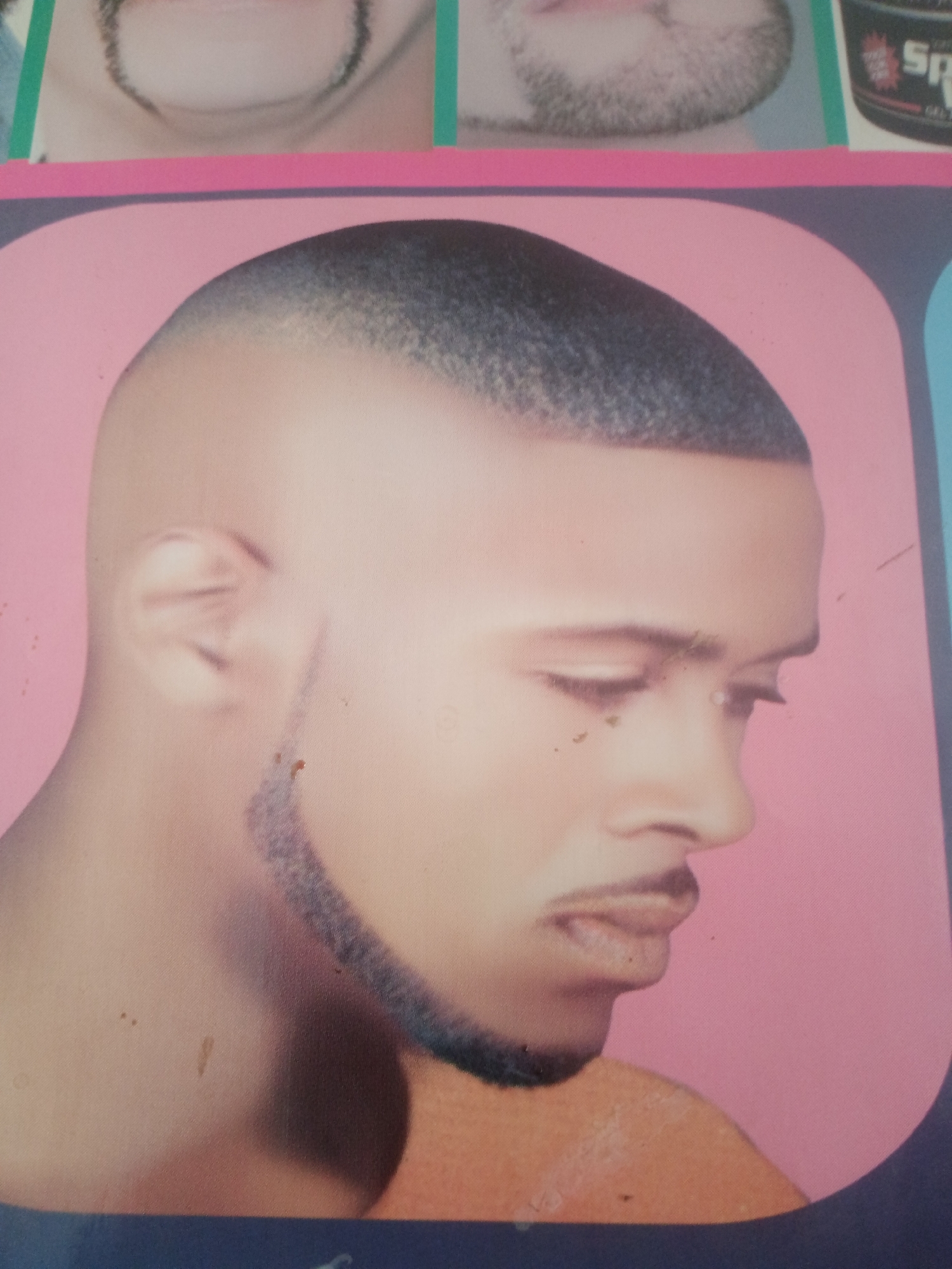 This is a photograph of a man's face, cropped to show his head and shoulders. He has a faded haircut, and his facial hair includes a mustache and a goatee. The image is set against a pink background, and appears to be a poster or advertisement for a barbershop. There are no identifiable landmarks or contextual clues within the image that would suggest its specific location in Jalingo, Nigeria. The image is also of low resolution and has some visual artifacts, possibly from printing or aging.