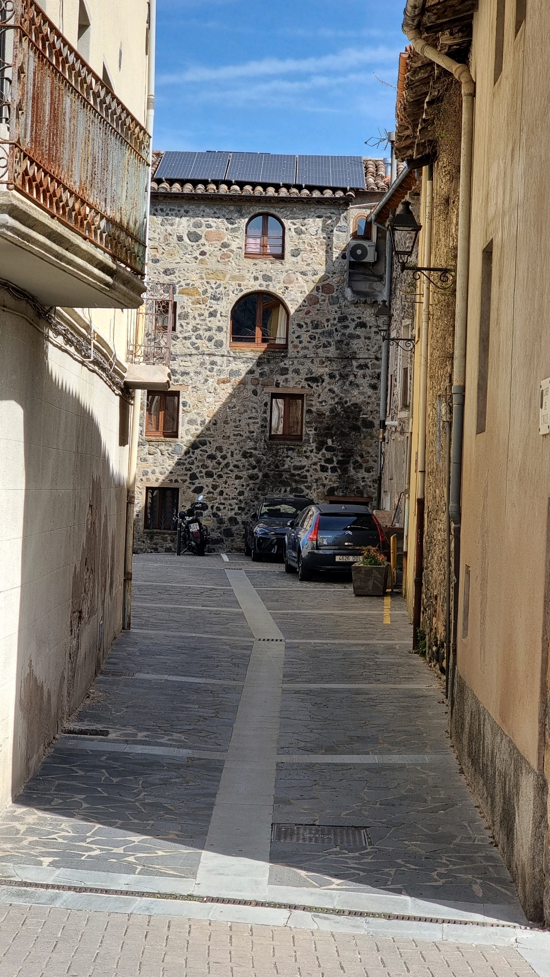 This is a vertical, eye-level, outdoor photograph taken in a narrow street in Castellfollit De La Roca, Spain. The scene is a street paved with irregular gray flagstones, bordered by buildings on both sides. The left side of the image features a light-colored building with a rusted metal balcony and a drainpipe. The right side of the image is dominated by a building with a textured, light brown facade, featuring a drainpipe and two vintage-style street lamps.
In the background, a stone building with a tiled roof is visible. This building has solar panels installed on its roof. It has several windows, some of which are arched, with wooden frames and shutters. An air conditioning unit is mounted on the exterior wall of this building.
Parked on the street are two dark-colored cars and a black motorcycle. The rear of a dark blue hatchback car with license plate "4626 DOL" is prominent in the foreground on the right. A small planter box is situated near this car. The street is partially covered in sunlight and shadow, creating a dynamic pattern. The sky above is blue with scattered white clouds.