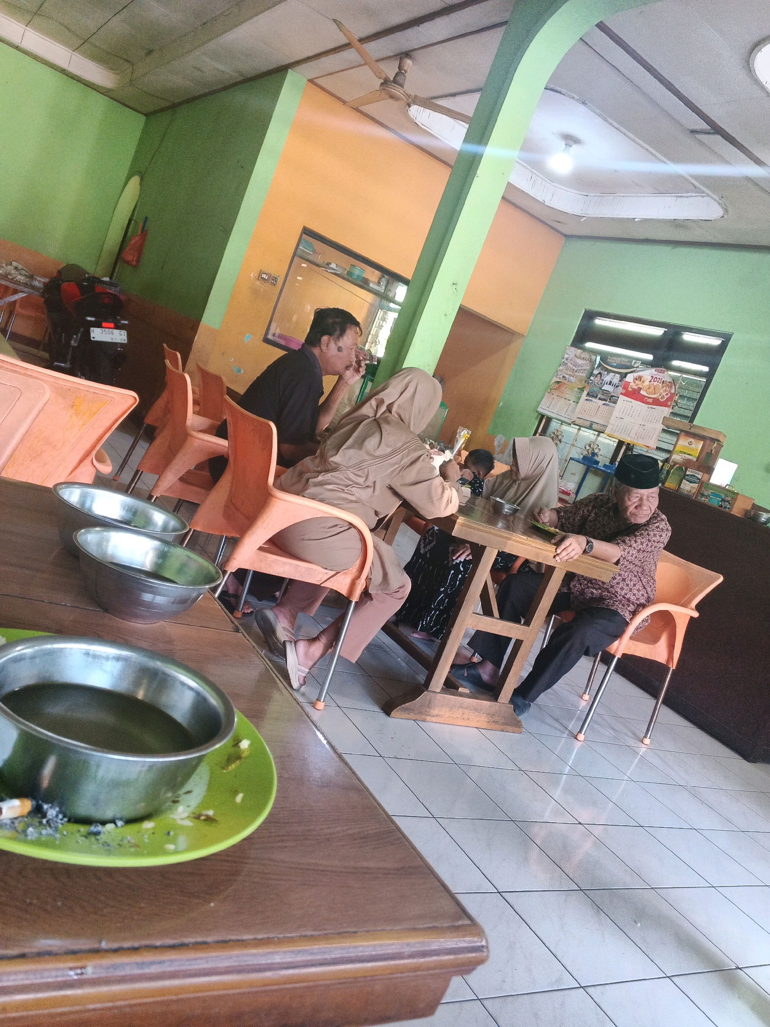 The image depicts a scene inside a casual eatery in Ungaran, Indonesia. The environment is characterized by bright, contrasting wall colors: lime green and orange. Several individuals are seated around tables, engaged in conversation or eating. In the foreground, a wooden table holds two stainless steel bowls, one of which contains a dark liquid and is placed on a green saucer with a lit cigarette and ash. To the right, an elderly man wearing a patterned shirt and a peci hat is seated at a wooden table, smiling and looking to his left. Two women, one wearing a beige hijab and the other a tan hijab and dress, are also seated at tables, one of them appearing to interact with the elderly man. In the background, a motorcycle is parked near an orange chair. The floor is tiled with white squares. The overall atmosphere suggests a relaxed, everyday gathering.