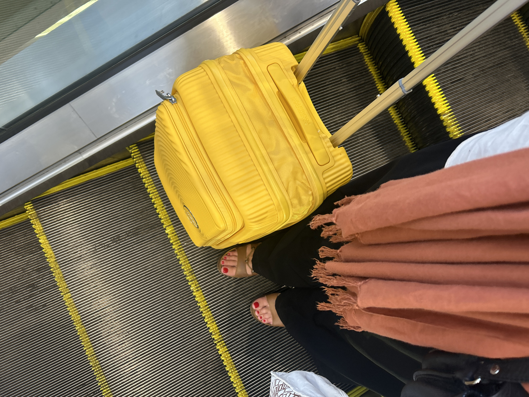 An overhead shot shows a person on an escalator in Mai Khao, Thailand, with a bright yellow suitcase. The escalator steps are dark gray with yellow safety lines. The person is wearing black pants and tan sandals with red nail polish. A coral-colored scarf is draped over their lap, and a white bag is partially visible on the left. The suitcase, positioned to the right of the person, is a large, hard-shell model with wheels and an extended handle. The lighting suggests it is daytime.