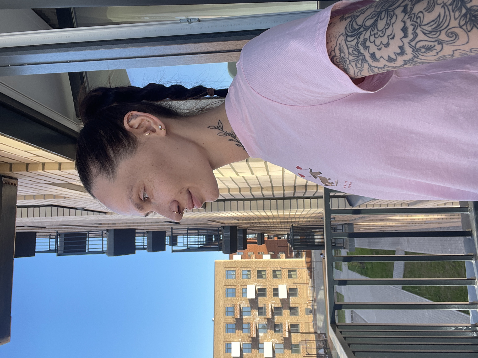 A medium shot depicts a fair-skinned person wearing a pink t-shirt, standing on a balcony. The individual has dark hair pulled back into a braid and several ear piercings. A tattoo is visible on the upper left arm and the neck.
The balcony features a dark metal railing. The backdrop shows a cityscape on a sunny day with clear blue sky. Two buildings are visible, one brick and one tan, with several windows. A patch of greenery is seen between the buildings. Based on visual clues (architecture), this location is possibly in the city of Izhevsk, Russia.