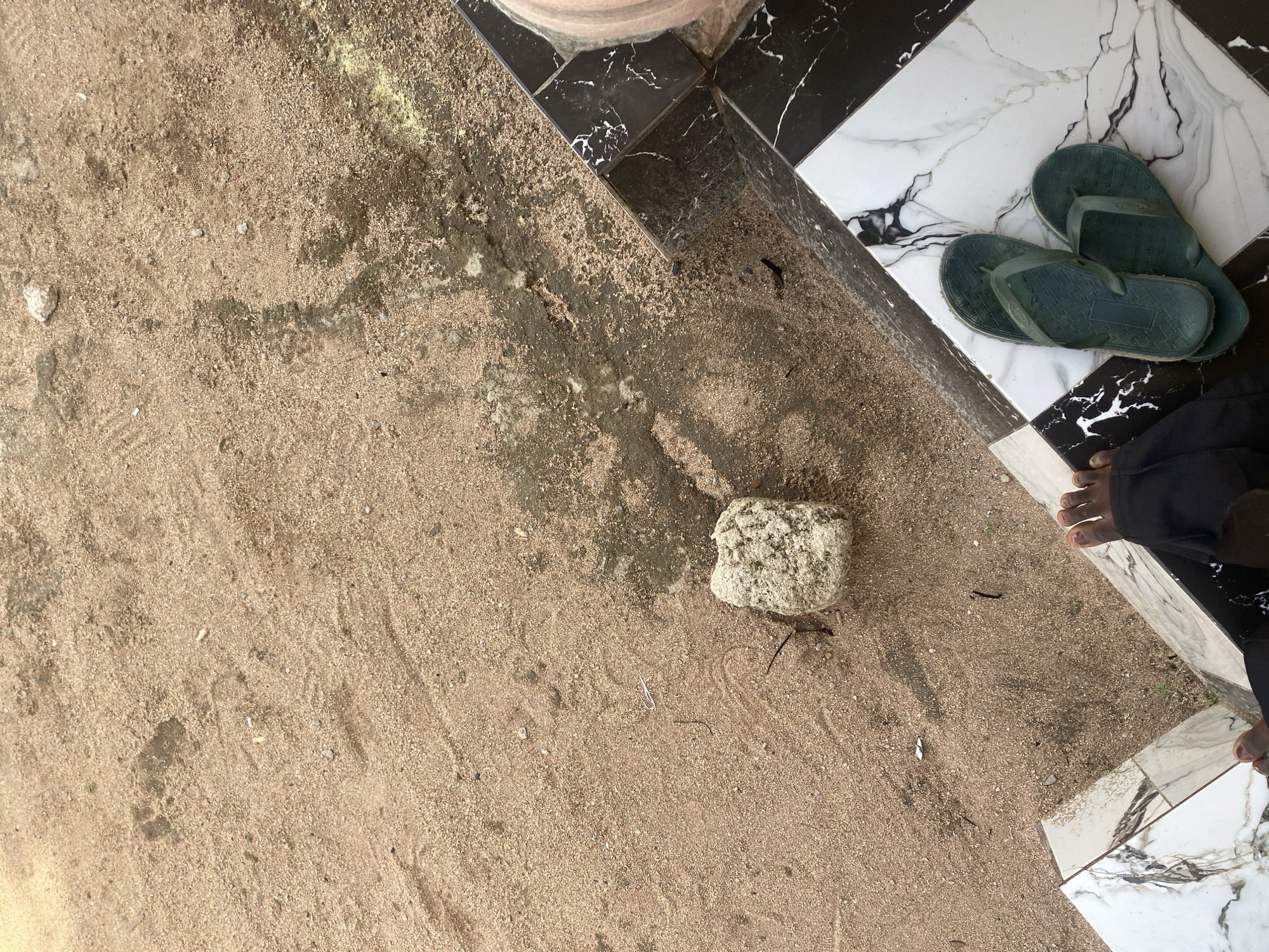 A pair of green flip-flops rests on a patterned tiled floor, which is a mix of black, white, and grey marble-like squares. To the left of the tile, a sandy area is visible. This sandy area has some debris, including a light-colored, rough-textured rock, and faint impressions that could be footprints. The scene appears to be an outdoor location, possibly near an entrance or walkway in Jauro Yinu, Nigeria.