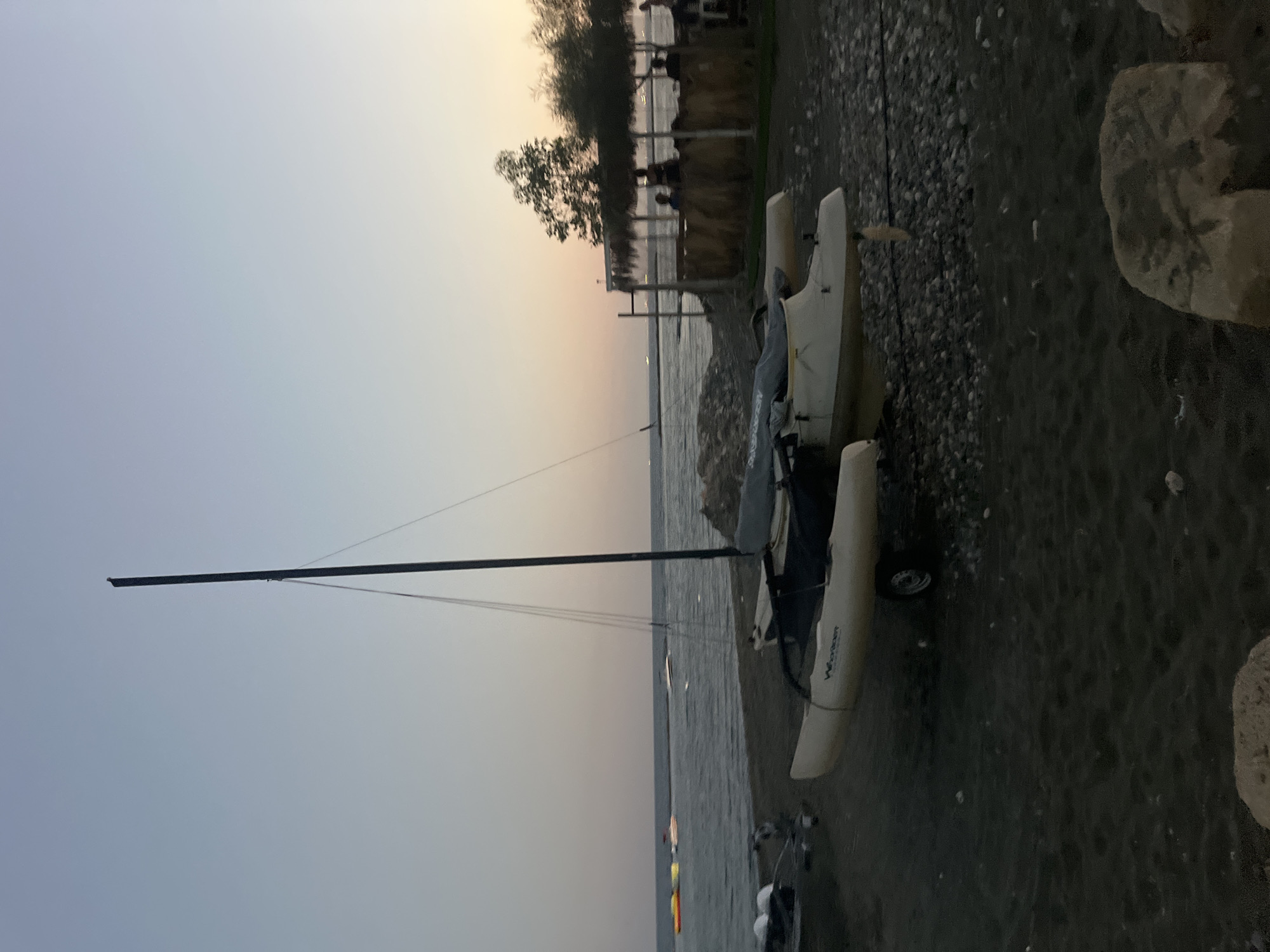 A catamaran sailboat is beached on a dark sand and pebble shore in Agios Tychona Community, Cyprus, at dusk. The sky transitions from a pale blue overhead to a soft pink and orange near the horizon, indicating the sun has recently set. The calm sea stretches out in the background, with a few other boats and water toys visible further offshore. To the right, a thatched-roof beach bar structure with visible patrons is situated behind a wooden barrier. The catamaran, a light-colored two-hulled vessel with a tall mast, is resting on a trailer. Several other smaller boats and watercraft are also visible on the shore in the foreground and midground. Rocks are scattered across the beach.
