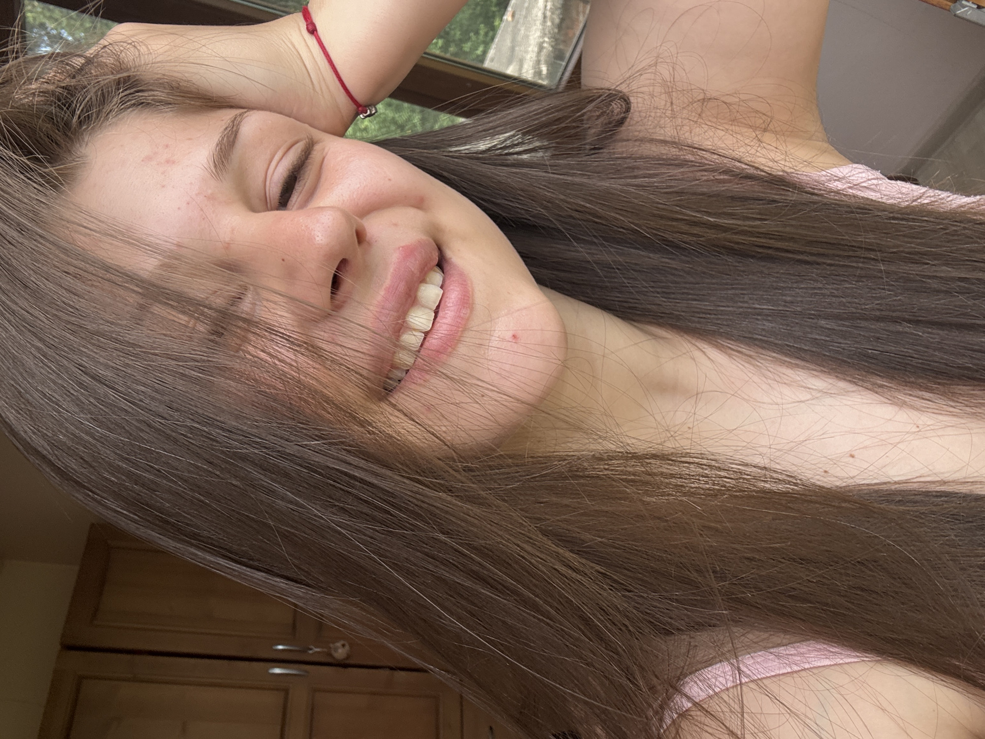 A close-up, slightly angled, medium shot captures a young woman with long, brown hair covering part of her face. She is smiling with her eyes partially closed, her right hand resting on her head. She wears a pink camisole, and a red string bracelet is visible on her wrist. The background shows wooden cabinetry, a window with greenery outside, and a wall. The lighting suggests an indoor setting with natural light coming from the window.
