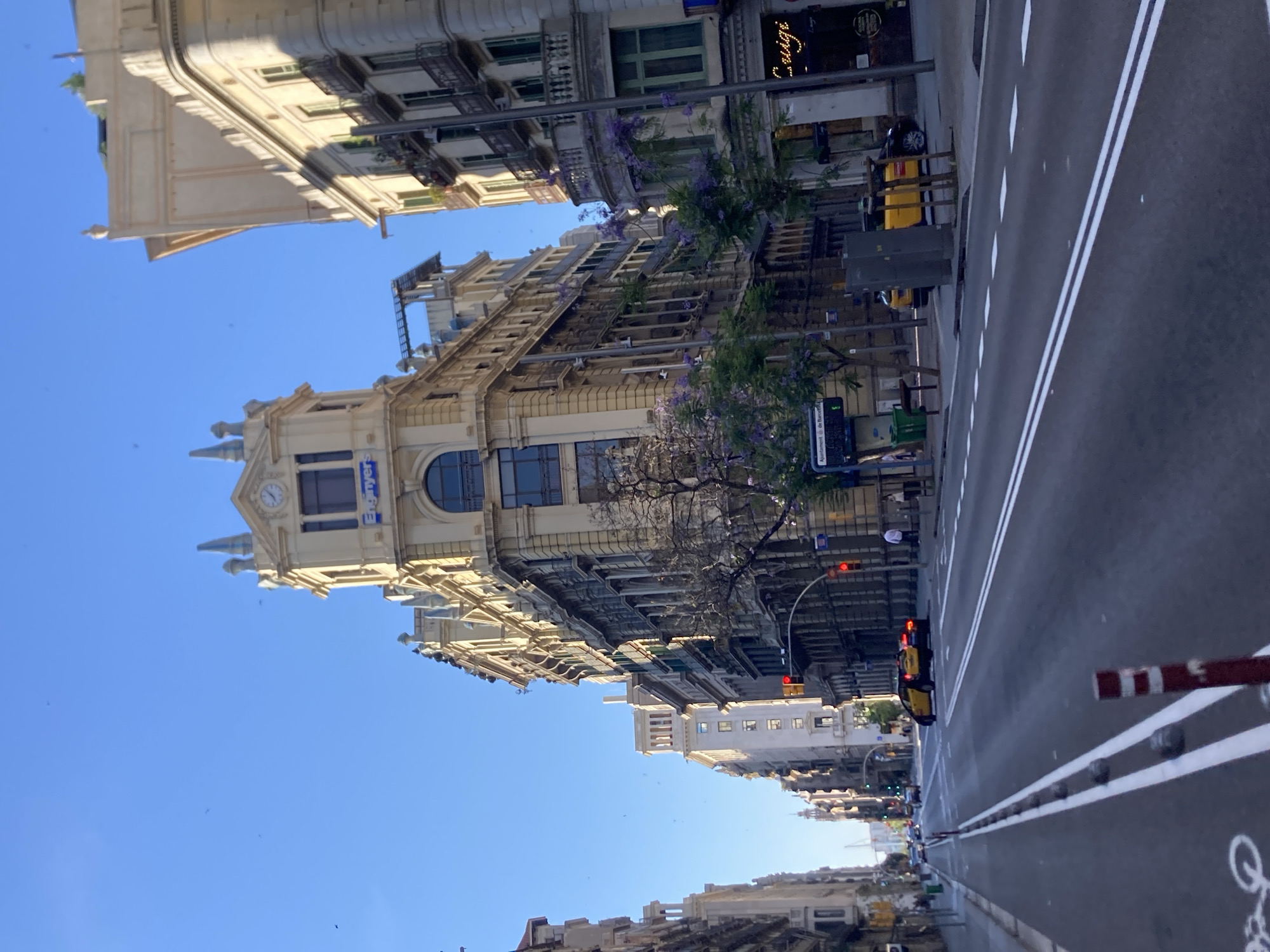 This image captures a street scene in Barcelona, Spain, likely on a sunny day given the bright blue sky and strong shadows. The perspective is from street level, looking down a wide, multi-lane road lined with grand, old European-style buildings. The architecture is characterized by ornate facades, balconies, and decorative upper levels, with a prominent building in the foreground featuring a clock tower and a sign that reads "Enginyers".
The road itself has distinct lane markings, including white lines and a dedicated bike lane indicated by white paint. Several cars are visible, including two yellow taxis, one of which is in motion. Pedestrians are present on the sidewalks, though they are small in the frame. Trees with purple blossoms are planted along the sidewalk, adding a touch of color to the urban landscape. Traffic lights are visible, indicating an intersection ahead. The overall impression is of a vibrant, active city street with a rich architectural history.
