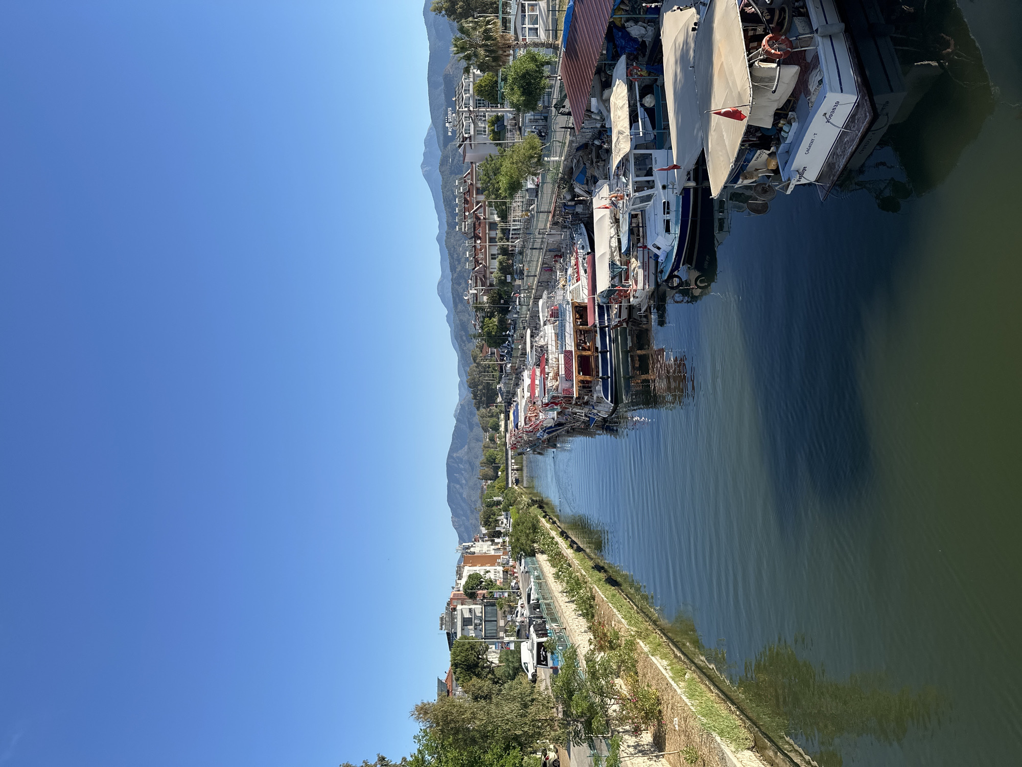 The image depicts a waterfront scene in Fethiye, Turkey. A canal or river is lined with numerous boats docked closely together on one side. A pedestrian walkway runs along the opposite bank, separated from the water by a low stone wall with some vegetation. Beyond the walkway, there are buildings and parked vehicles. In the background, mountains rise above the town. The sky is clear and blue.