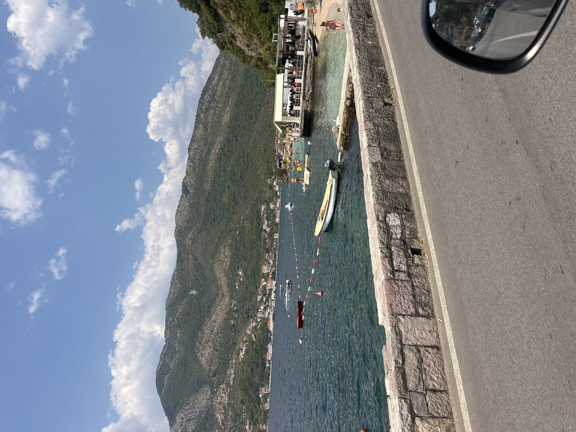 In Lepetane, Montenegro, a sun-drenched coastal scene unfolds under a bright blue sky with scattered white clouds. A stone wall separates a paved road from the shimmering blue sea. Bouys and ropes delineate a swimming area in the water. A red boat and a light-colored dinghy are moored near the shore. A beach area is visible in the distance, with a white-canopied structure resembling a restaurant or cafe. Several people are present on the beach and in the water, some swimming and others gathered at the establishment. A forested mountain range rises in the background, with scattered buildings dotting its slopes. A car's side mirror is visible in the lower right corner of the frame, reflecting a road and parked vehicles.