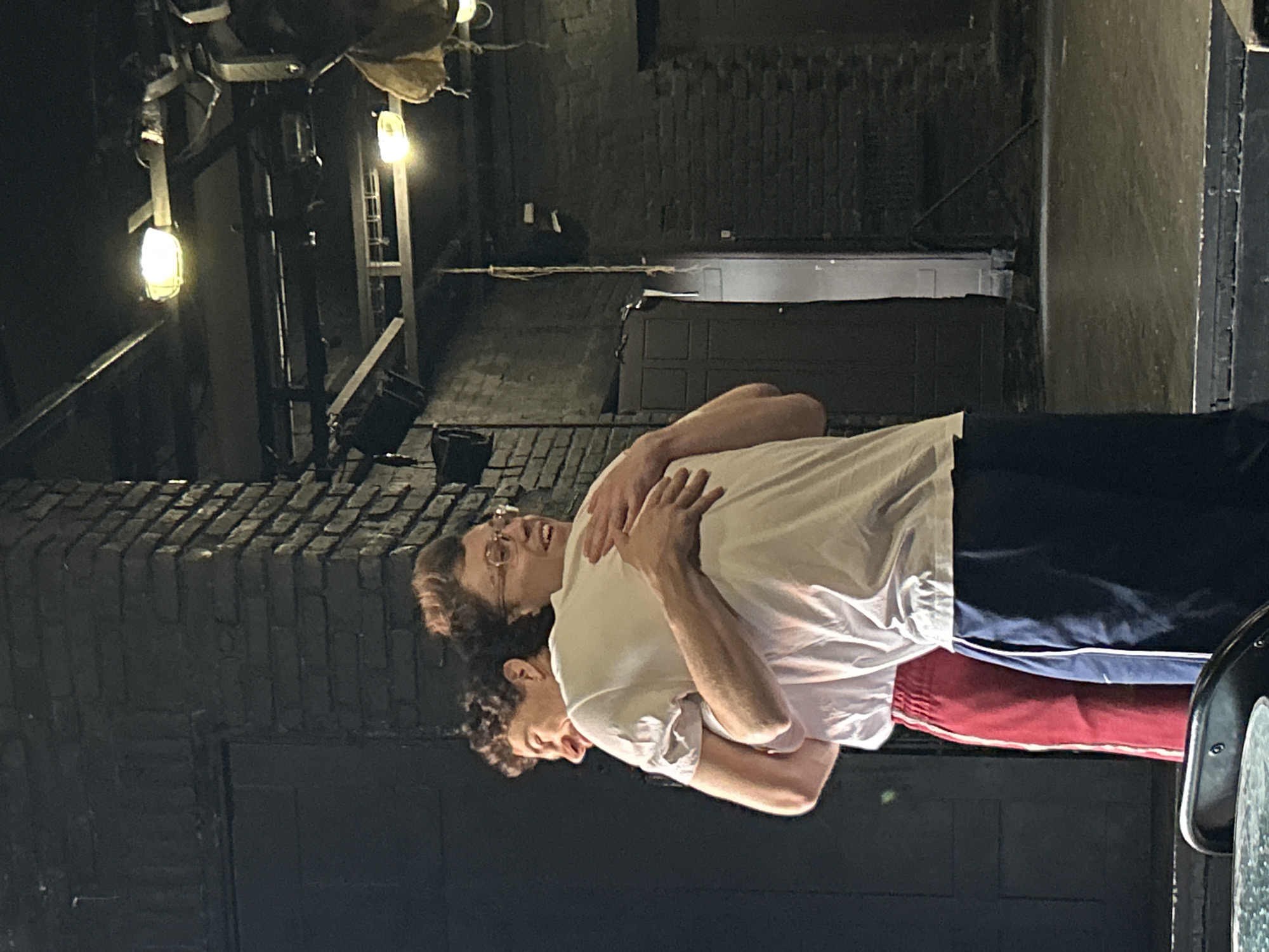 Two people are embracing on a stage. The person in the foreground, wearing glasses and a white t-shirt, is hugging the other person from behind. The person being hugged is wearing a white t-shirt and red athletic pants with a white stripe. The background consists of a brick wall, dark doors, and theatrical lighting fixtures. The stage floor is visible and appears to be made of wood. The overall scene suggests a moment of affection or camaraderie between the two individuals.