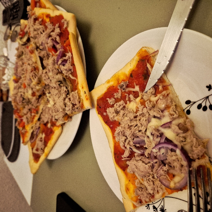 Two plates each contain a slice of homemade pizza with thick crusts. The pizzas have a red sauce base, topped with shredded tuna and thin rings of red onion. A serrated knife cuts into the pizza on the right plate, near a fork. One plate features a black and white design, while the other is plain white. The background appears to be a muted green surface.
