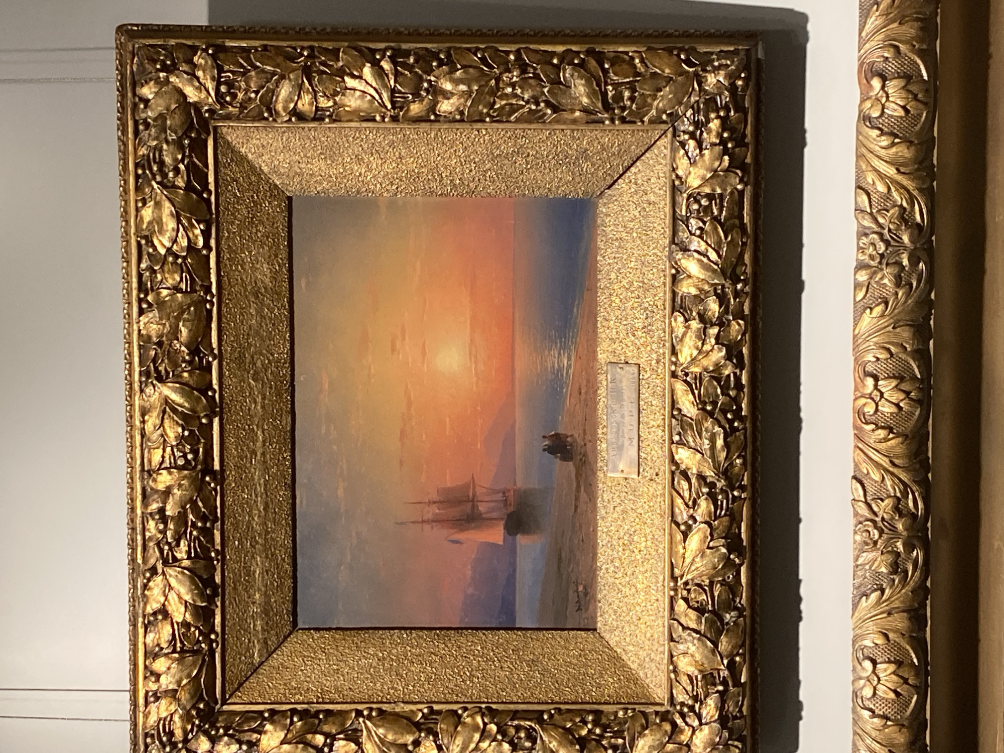 A framed painting hangs against a white wall. The painting depicts a sunset scene at sea with a tall ship silhouetted against the light. Two figures stand on the shore. The frame is ornate and gilded. A second, different gilded frame is partially visible to the right. The location is likely a museum in Yekaterinburg, Russia.
