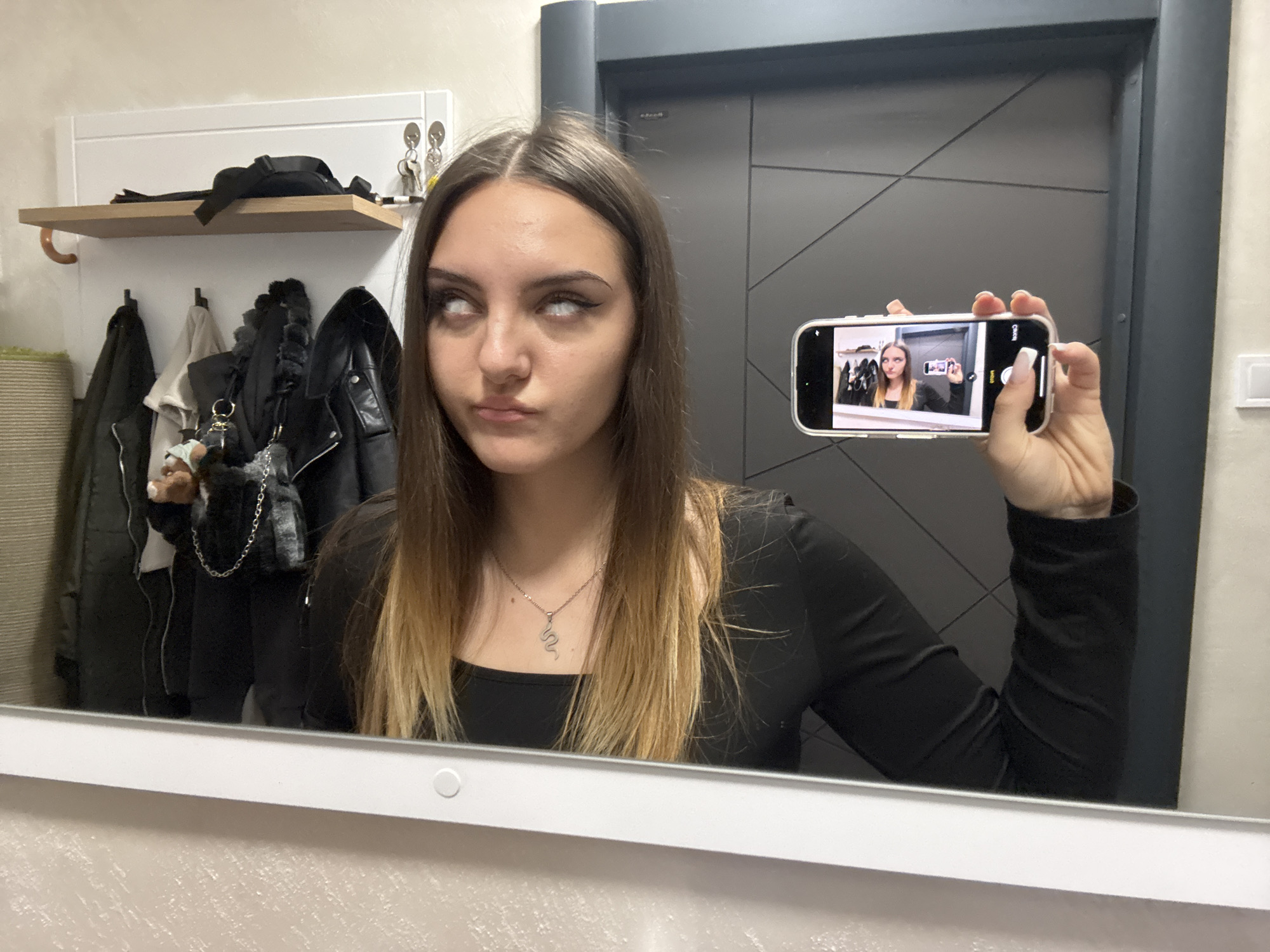 A woman is reflected in a mirror, holding a smartphone showing a smaller image of herself in the same pose, creating a recursive effect. The woman in the reflection has rolled her eyes back, showing only the whites. She has fair skin, dark eyeliner, and long brown hair with blonde highlights. She wears a black long-sleeved top and a silver snake pendant necklace. Behind her are a closed gray door and a white shelf with clothes and keys. The mirror's frame is off-white. The location is likely a bathroom, as suggested by the mirror and the coat rack in the background. The geographical location of Sofia, Bulgaria cannot be determined from this image alone.