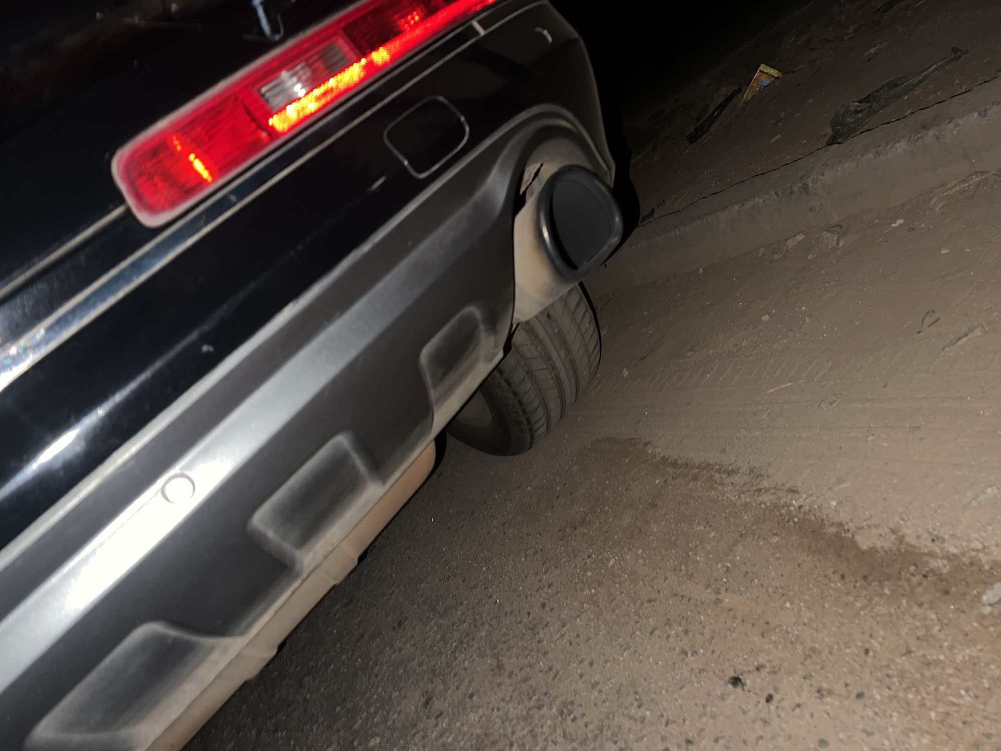 The image shows the rear of a black car parked on the side of a paved road, possibly in Kubwa, Nigeria. The car's taillight is illuminated. The road surface appears to be worn. There is a patch of debris or litter along the roadside.