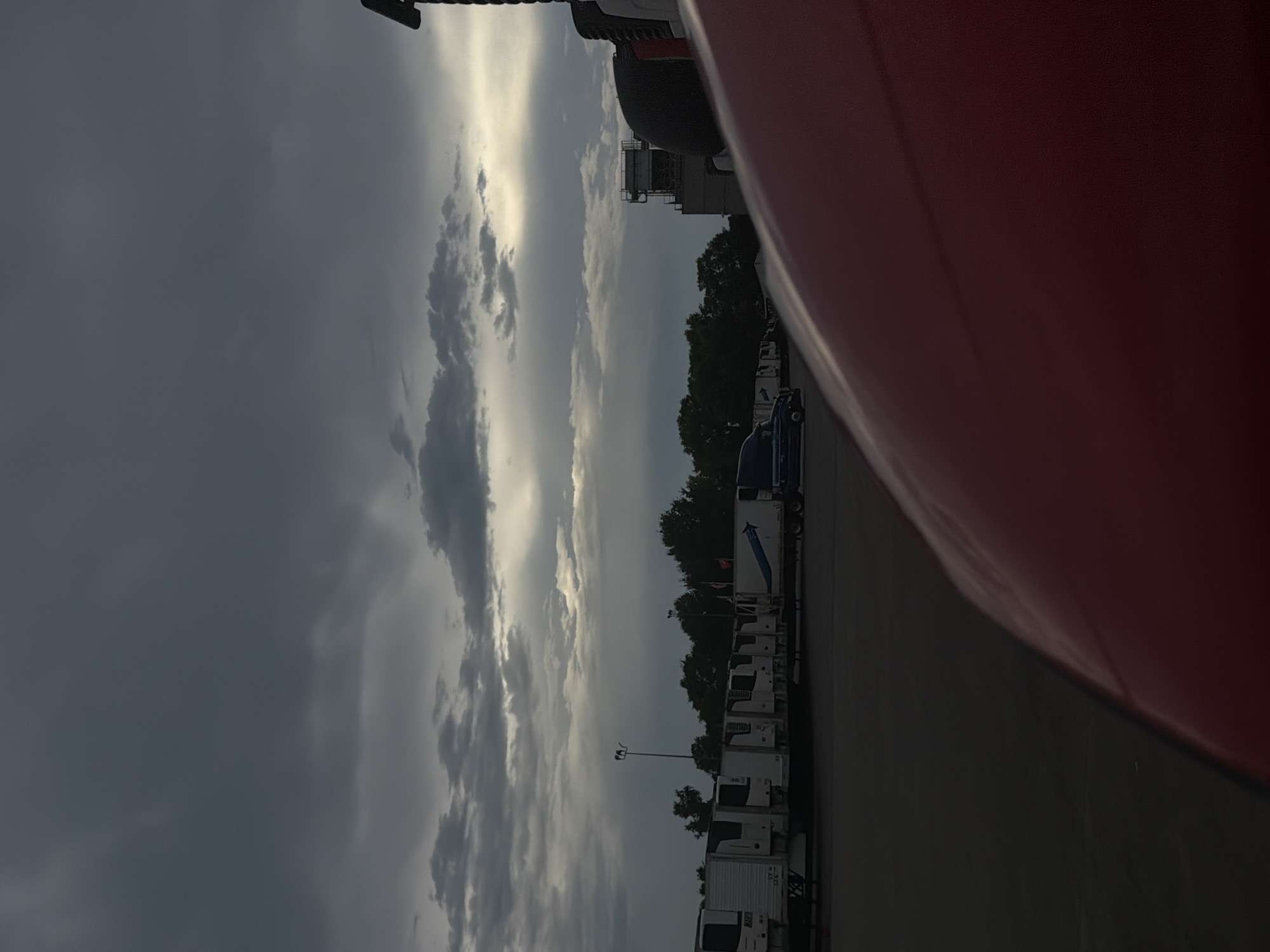 The image depicts a parking lot with several semi-trailer trucks and a building in the background under a cloudy sky. The foreground is dominated by the red hood of a vehicle. In the middle ground, a row of refrigerated trailers is parked, some bearing the brand "MARK." A blue semi-trailer truck is also visible, parked to the left of the trailers. The background features a line of trees and a multi-story building with industrial-looking structures. The sky is filled with dark, dramatic clouds, with a sliver of sunlight breaking through on the right side. The scene suggests an overcast or approaching storm. Based on the metadata, the location is Murfreesboro, United States of America.