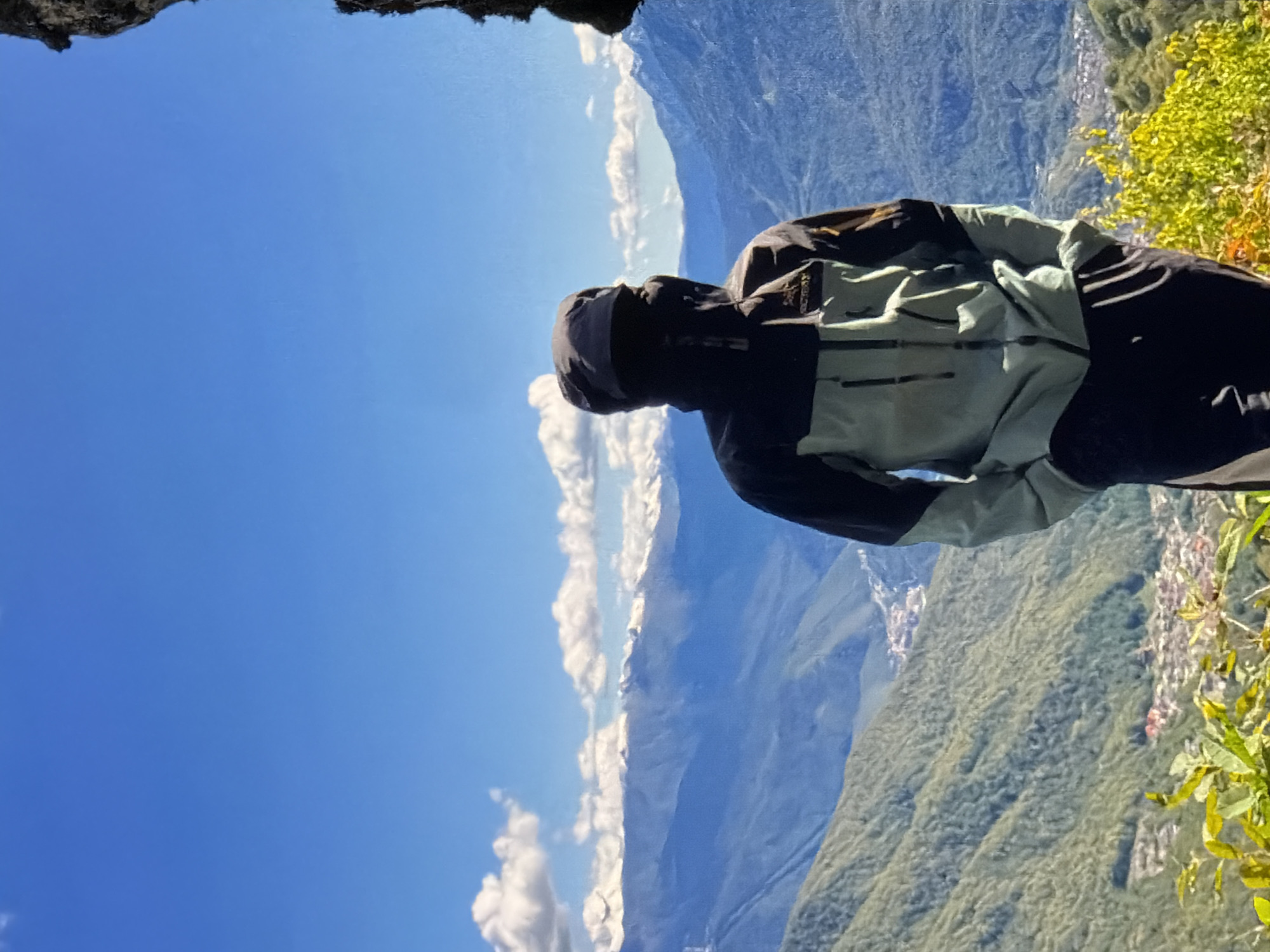 A person in a black and light blue jacket stands on a mountain overlooking a valley with a clear blue sky and some white clouds in Unknown, Unknown. The foreground shows some green vegetation, and in the distance, there are mountains and a small town.