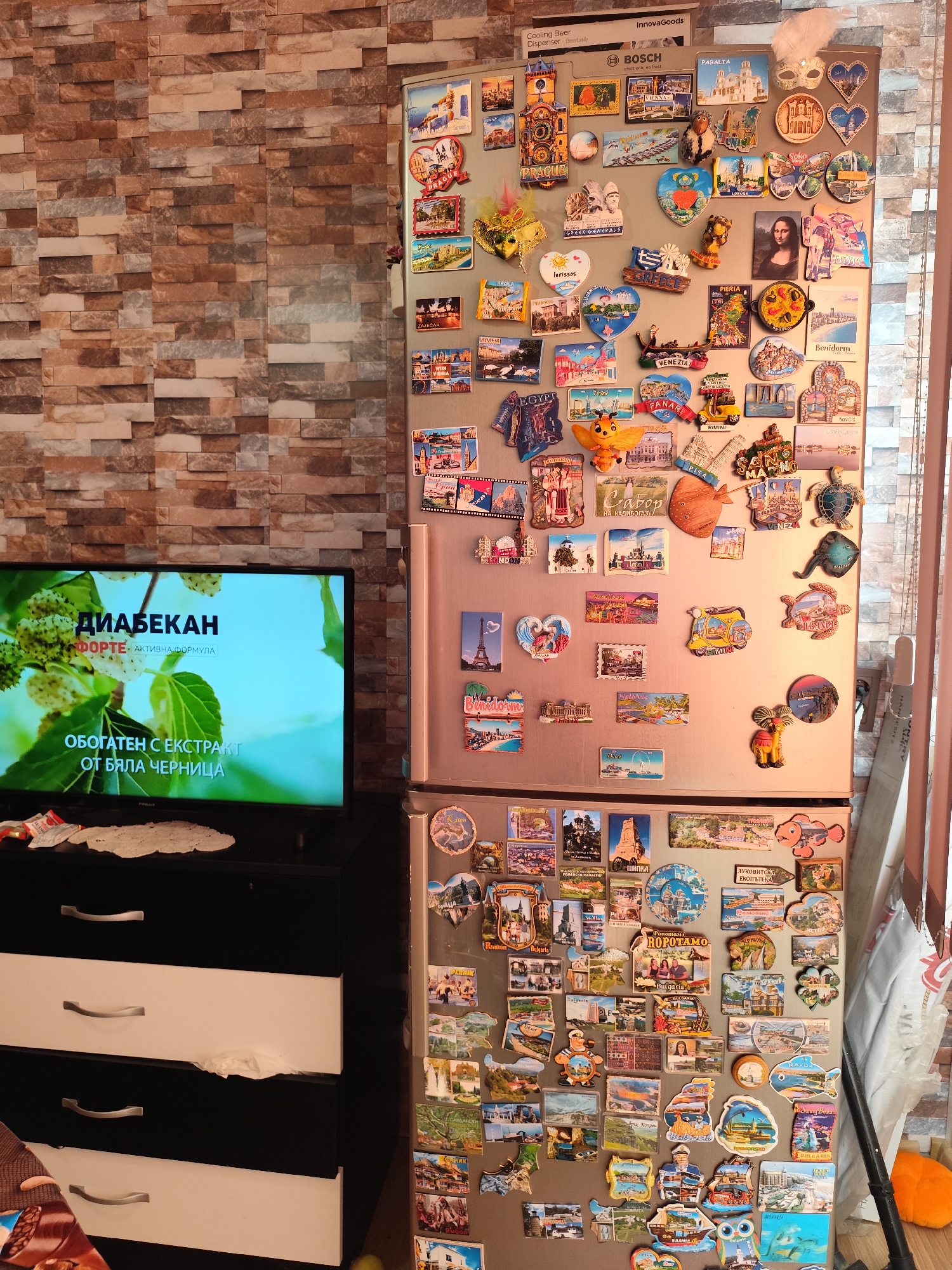 The image shows a refrigerator heavily adorned with numerous colorful magnets, predominantly depicting tourist attractions and landmarks from various locations. The magnets feature images of cities, historical sites, and scenic landscapes, suggesting a collection of souvenirs from travels. Some magnets are shaped like iconic objects, such as masks or vehicles. The refrigerator itself is a standard appliance, with a brand name "BOSCH" visible at the top. To the left of the refrigerator, a television screen displays an advertisement with text in Bulgarian, specifically mentioning "ДИАБЕКАН" and "БЯЛА ЧЕРНИЦА" (White Mulberry), which is likely a health-related product. The backdrop is a wall with a textured, brick-like pattern, suggesting an indoor domestic setting. The overall impression is that of a lived-in space with personal mementos displayed. The presence of the Bulgarian text on the television strongly suggests the location is in Bulgaria. While the image is not specific to the city of Montana, Bulgaria, it is consistent with a Bulgarian household.