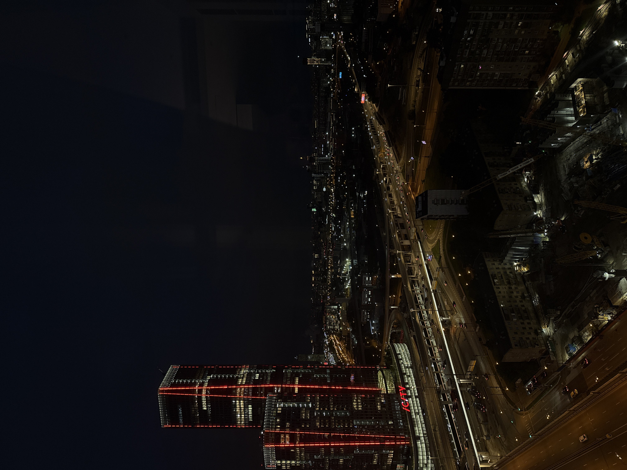 The image depicts an aerial view of Khimki, Russia, at night. The lower portion of the frame features a modern high-rise building with distinct horizontal red light strips. The word "CITY" is vertically spelled out in red on one side of the structure. Adjacent to the building are transportation infrastructures, including roads, overpasses, and what appears to be train tracks. Further into the image, a cityscape stretches into the distance, with various lights from buildings and street lamps illuminating the darkness. The upper left corner of the image consists of a dark, undefined space, possibly representing the sky or a shadowy foreground.