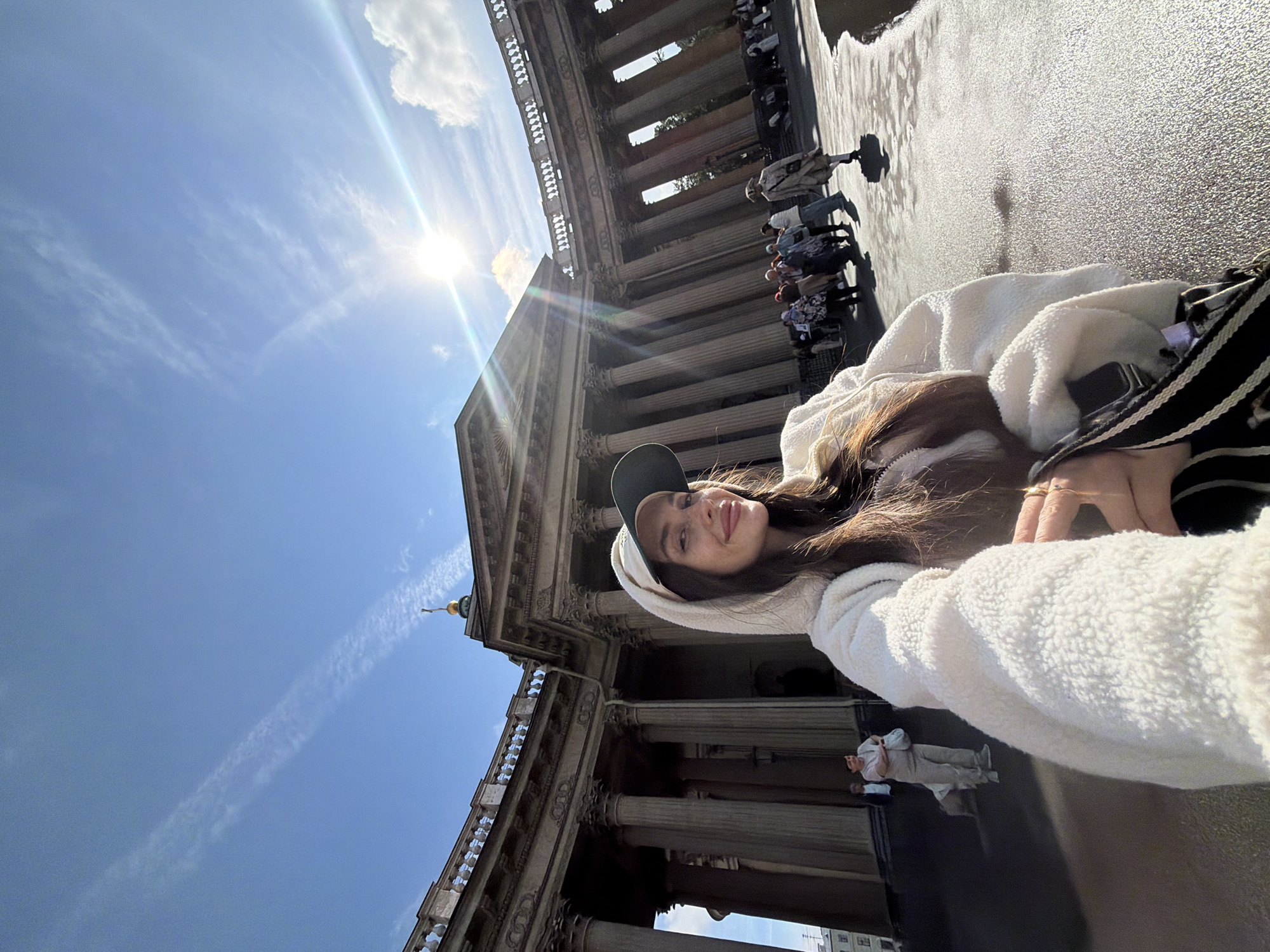 A woman in a white hoodie and a baseball cap takes a selfie in front of Kazan Cathedral in Saint Petersburg, Russia. The sun shines brightly in the blue sky, with some wispy clouds. People can be seen walking around the cathedral, which is a large, neoclassical building with a semi-circular colonnade. The woman is smiling and looking at the camera, holding a bag in her hand. The overall impression is of a sunny day in a historic city.