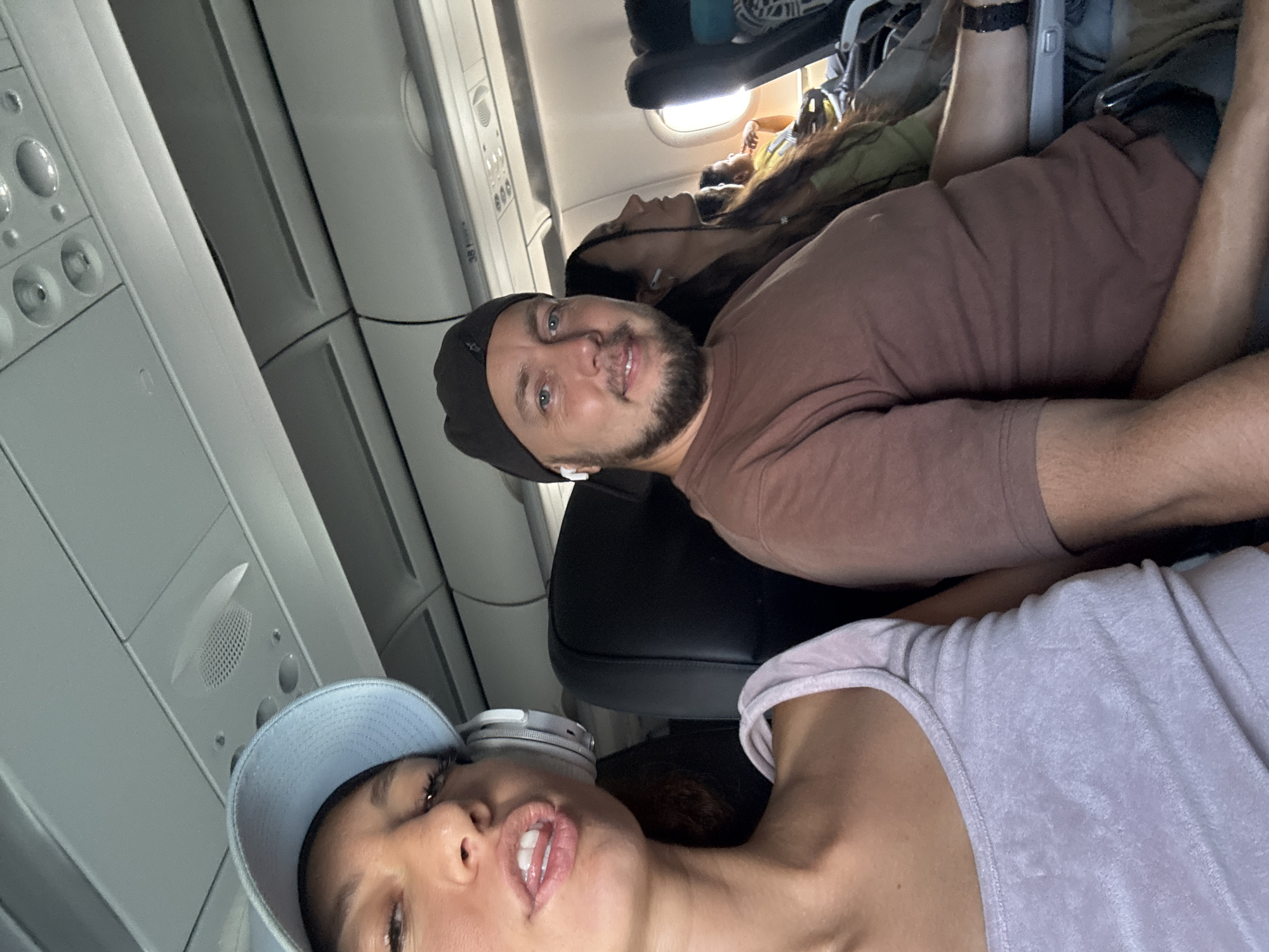 Onboard an airplane, a selfie is taken. Three people are visible: a woman in a light purple shirt and blue baseball cap, a man in a brown shirt and black beanie, and another woman lying behind the man. The airplane's interior is visible, including overhead compartments, air vents, and seats. The location is not specified.