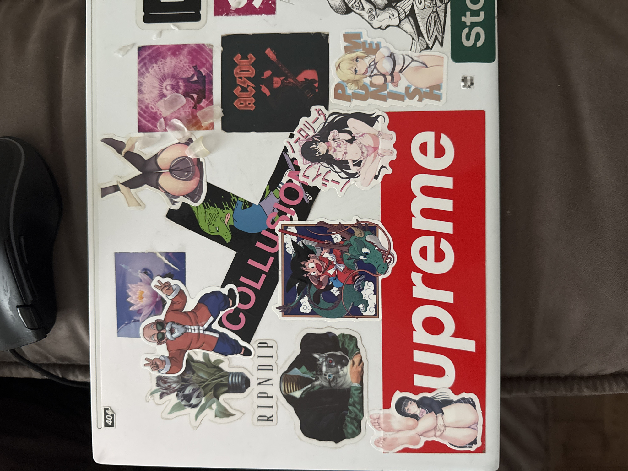 The image shows a laptop covered in various stickers. The stickers depict anime characters, a cat wearing a suit, a lotus flower, the band AC/DC, and the brand Supreme. There is also a sticker with the text "RIPNDIP". The laptop is resting on a dark, textured surface, likely a couch or blanket. The background is dark and out of focus. The location in the city of Бараўлянскі сельскі Савет, Belarus, is not determinable from the image.