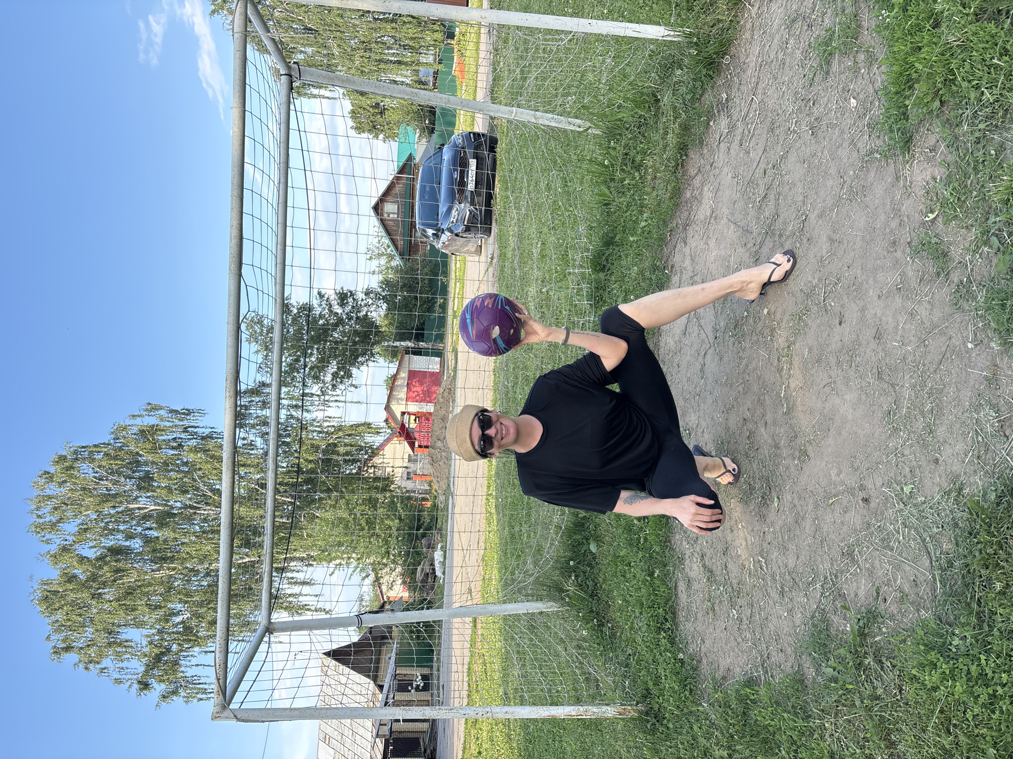 A woman wearing a black t-shirt, black leggings, a hat, and sunglasses is squatting in front of a soccer goal. She is holding a purple and blue soccer ball in her right hand. The soccer goal is made of metal and has a net. The background shows a grassy field, trees, houses, and a black car. The location is likely a rural or suburban area in Chaikovsky, Russia, given the architecture and vegetation.
