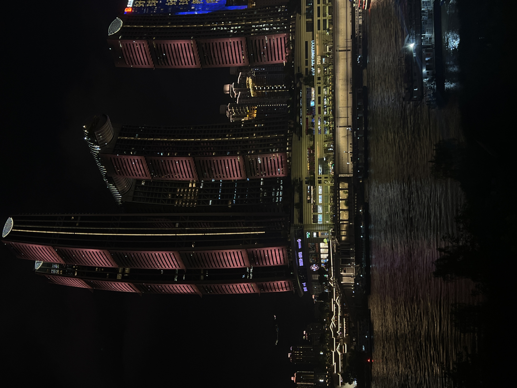 The image captures a night view of the Chongqing Guanyin Bridge Financial District in Jiangbei City Street, China. The scene is dominated by illuminated skyscrapers, with the most prominent featuring distinctive pink lighting on their facades, interspersed with vertical golden lights. One of the skyscrapers has a unique architectural design with curved elements at its top. In the foreground, a wide river flows, reflecting the city lights. Several boats and barges are visible on the water. Along the riverbank, a multi-story building complex is brightly lit with various signs and illuminated windows. Further in the background, other buildings of varying heights are visible, also illuminated against the dark night sky. The overall impression is one of a vibrant, modern urban landscape at night.