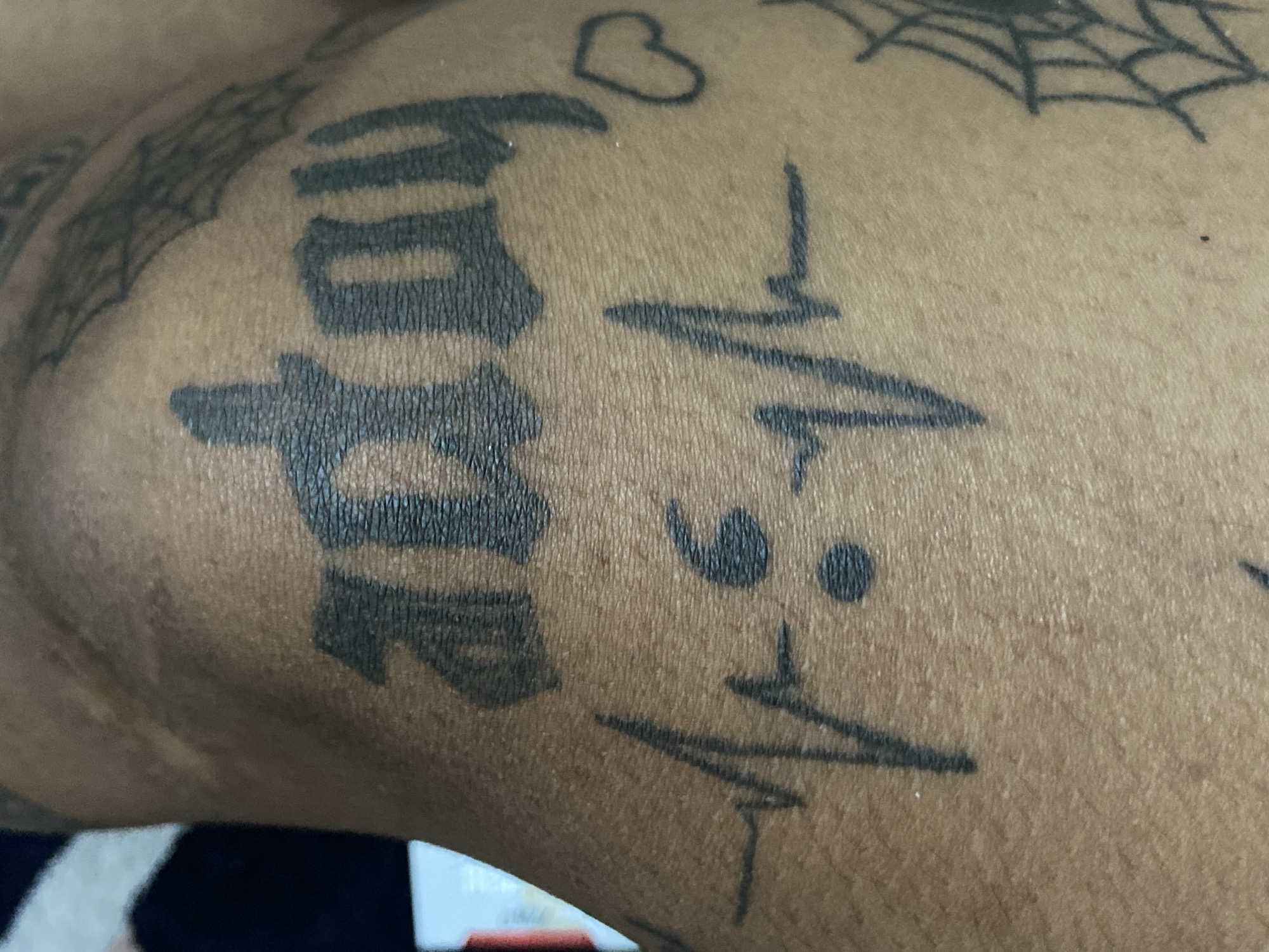 This is a close-up image of a person's arm or shoulder, showcasing several tattoos on a medium to dark skin tone. The skin shows visible texture, suggesting a detailed shot.
The most prominent tattoo features the word "HOPE" in a bold, block-like font, oriented vertically down the arm. Above the "H" in "HOPE," a small outline of a heart is visible. To the left and slightly above the "HOPE" tattoo, parts of a spiderweb design are visible, with another section of a spiderweb appearing in the upper right corner of the frame.
To the right of the "HOPE" tattoo, there is a design resembling an EKG or heartbeat line. This line is interrupted by two vertically stacked dots, creating a symbolic element within the heartbeat pattern.
The background is softly blurred, showing indistinct shapes and colors, likely suggesting an indoor setting with no specific activity taking place. The tattoos appear established, not freshly applied. The given context for the location is Kuta, Indonesia.