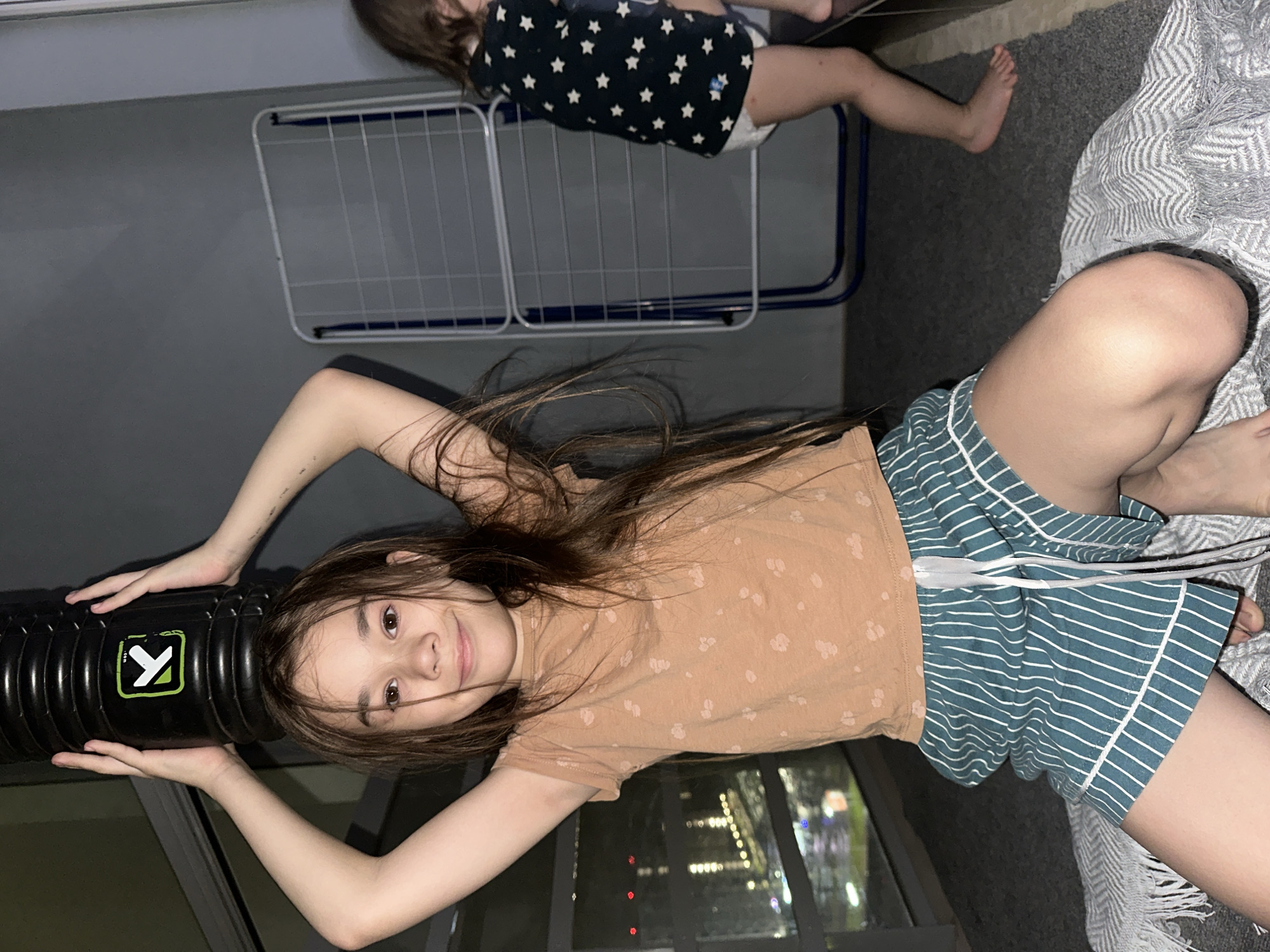 Two young girls are visible in what appears to be an indoor setting, possibly an apartment in Izhevsk, Russia, given the background light cues suggesting a nighttime city view.
The girl in the foreground, with long brown hair, is lying on her back, looking up at the camera with a slight smile. She is wearing a light brown short-sleeved top with small white patterns and teal and white striped shorts. Her arms are raised, holding a black foam roller with a green and white logo that resembles a stylized "X" or a checkmark. Her expression is calm and happy.
In the background, a second, younger girl is partially visible. She is wearing a dark top with white stars and what looks like a white diaper or pull-up. She is standing or crawling near a clothes drying rack, and her bare feet are visible on a dark gray carpet.
The overall scene suggests a relaxed, playful atmosphere, likely in the evening or at night, as indicated by the illuminated city lights visible through a window in the background. The interior includes a dark gray carpet and a gray wall or floor, with a textured gray blanket also partially visible on the right side of the frame. The clothes drying rack in the background is white with blue accents.
