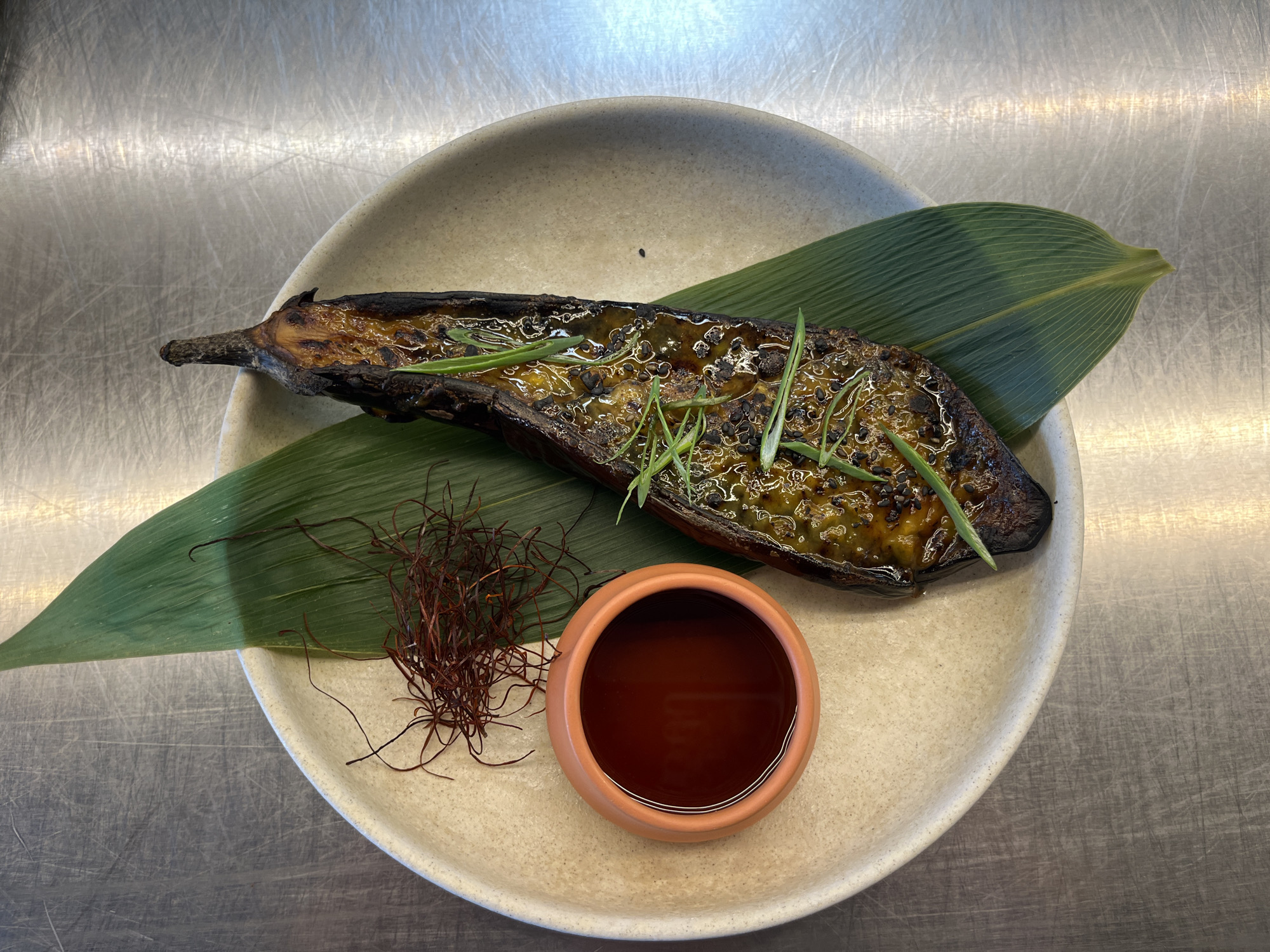 This image presents a gourmet dish meticulously arranged on a rustic plate, set against a backdrop of a scratched, metallic surface, typical of a professional kitchen or restaurant environment in Sofia, Bulgaria.
The central subject is a roasted eggplant, likely a Japanese eggplant, cut in half lengthwise. It is generously coated with a glistening, golden-brown glaze, possibly a sweet and savory sauce like miso. The eggplant is garnished with fresh, chopped green onions or chives and sprinkled with black sesame seeds, adding both visual appeal and textural contrast. The skin of the eggplant shows signs of charring, indicating a roasting or grilling process.
The eggplant is elegantly presented on two large, dark green leaves, which serve as a natural bed on the plate. To the left of the eggplant, there is a small mound of fine, dried red threads, which appear to be chili threads (gochugaru) or a similar spice, providing a vibrant color accent.
Adjacent to the chili threads, a small, terracotta-colored ceramic bowl is filled with a dark, reddish-brown liquid, likely a dipping sauce, broth, or dressing intended to accompany the dish.
The dish is served on a round plate with a light, speckled beige or off-white finish, contributing to a handmade or artisanal aesthetic. The lighting is bright and even, illuminating the food and its textures. There are no people visible, nor any discernible text. The scene captures a prepared dish ready for consumption, emphasizing culinary artistry and presentation.