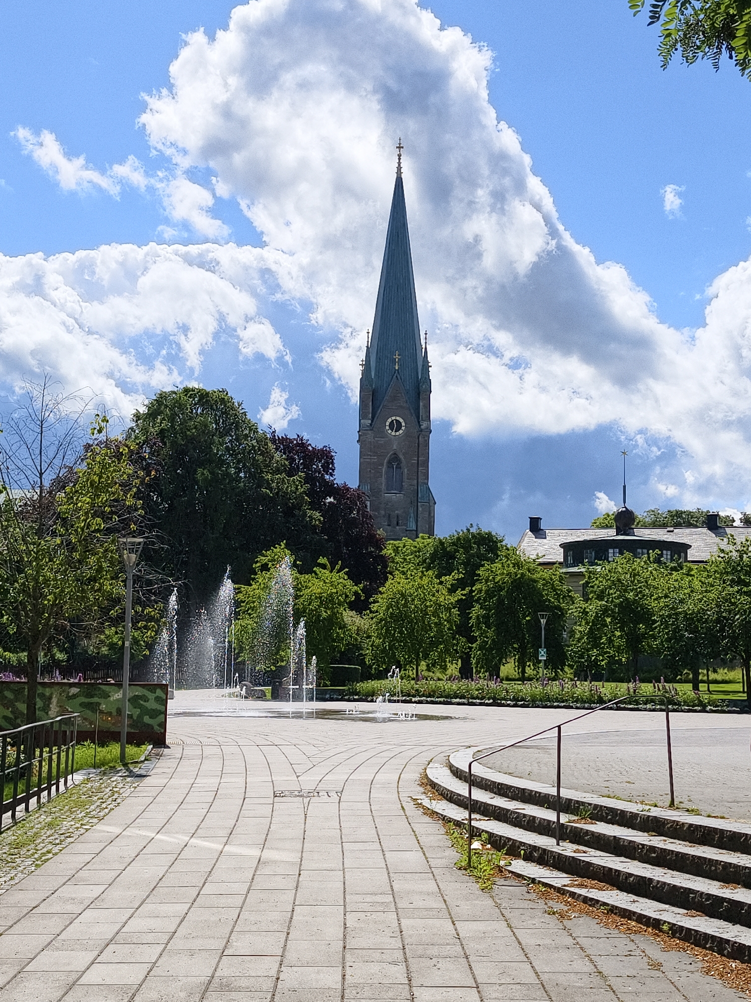 The image shows a public square with a fountain in the foreground and a tall church spire in the background. The square is paved with large, rectangular tiles, and there are several fountains spraying water. To the right of the square, there are steps leading up to a raised area. In the background, a church with a tall, slender spire is visible, along with trees and buildings. The sky is blue with scattered white clouds. The scene appears to be a sunny day in a city. This location is likely Trädgårdstorget in Linköping, Sweden, given the characteristic architecture of the cathedral.