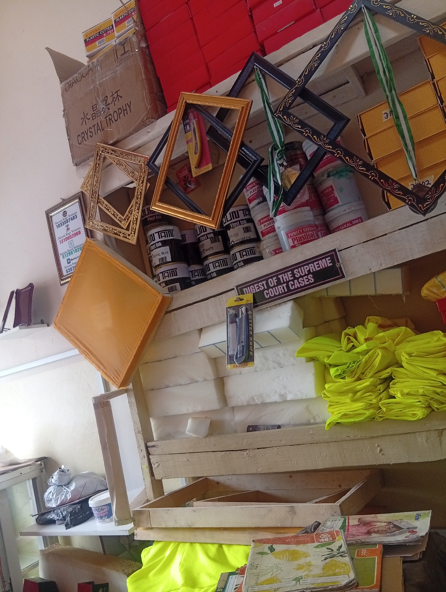 The image shows a cluttered storage area, likely in a small business or workshop. The scene includes numerous stacked and leaning items. On the top shelf, there are picture frames, ink containers, and boxes of plastic cutters labeled "KATENAI PLASTIC CUTTER K-701". There is also a cardboard box with Chinese characters and the English words "CRYSTAL TROPHY". On the middle shelves, there are white foam blocks, stacks of yellow fabric, and a "CUTTER KNIFE" package. The bottom shelf contains wooden frames and stacks of notebooks. A sign reads "DIGEST OF THE SUPREME COURT CASES". A business certificate for "Haruna Rogo Ventures" is visible on the wall. Given the business registration, the scene is most likely located in Sabon Gari East, Nigeria.