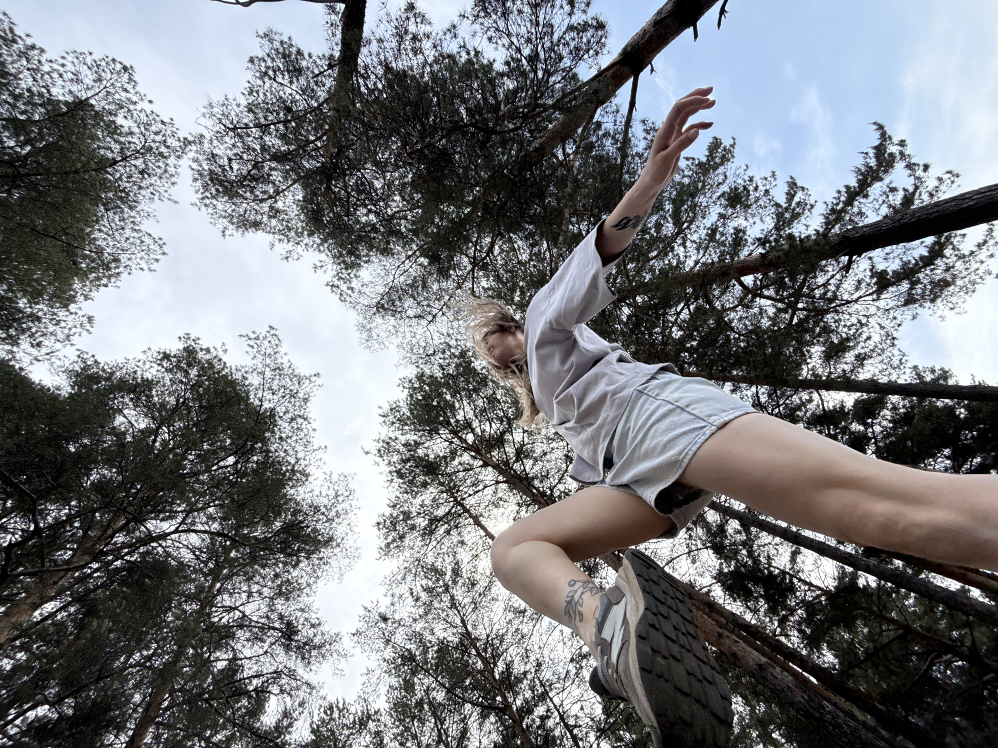 A low-angle, wide shot captures a person in mid-action, positioned against a backdrop of tall trees and a cloudy sky. The person appears to be stepping off a log or tree trunk, with their right leg extended forward and the left leg bent at the knee, foot still in contact with the surface. They wear light-colored shorts and a matching loose-fitting shirt. The person's left arm is outstretched, while the right arm is slightly bent at the elbow. Tattoos are visible on the person's left arm and right ankle.
The trees are tall and dense, forming a canopy overhead, obscuring a large portion of the sky. The sky is a mix of light blue and white clouds.
This is taking place in a wooded area in Chaikovsky, Russia.