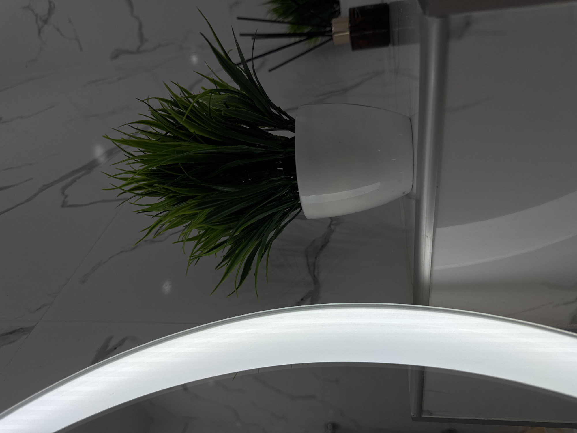 The image depicts a bathroom interior. A white pot with green, grass-like artificial plants sits on a counter. Beside the pot is a brown diffuser with black sticks. The background features a white marble-patterned wall. An illuminated, circular mirror is partially visible in the lower portion of the image. The location is inferred from the prompt, suggesting Stavropol, Russia.