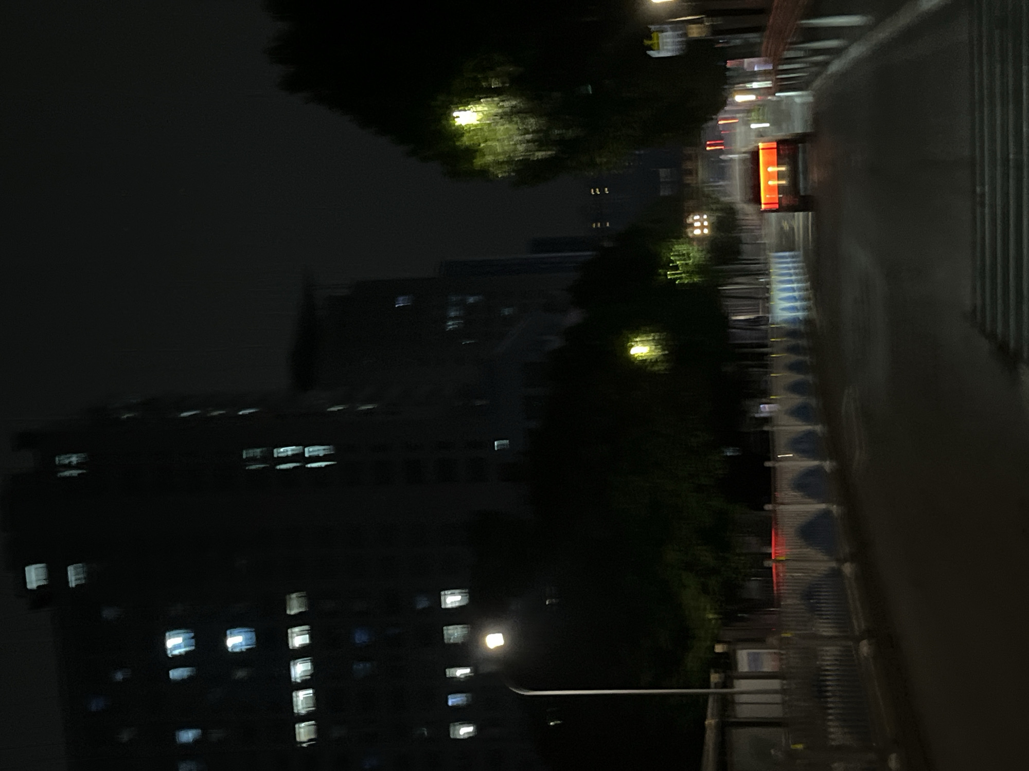 The image captures a nighttime scene on a street in Yuzhong District, China. Tall, dark buildings with illuminated windows line the left side of the street. A street lamp with a bright light is visible in the foreground on the left. Trees with blurred foliage are on the right, some illuminated by yellow streetlights. The road appears wet, reflecting lights. In the distance, a car with bright orange taillights is driving away from the viewer. A pedestrian crossing is visible in the foreground. The overall atmosphere is dark and somewhat blurry, suggesting it may have been taken from a moving vehicle or in low light conditions.