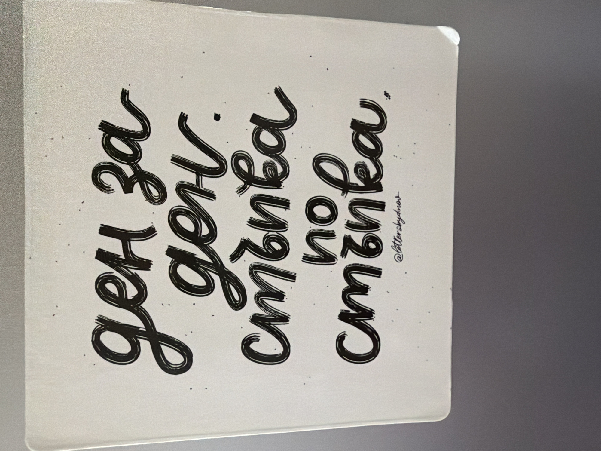 The image is a close-up, eye-level, vertical shot of a white, rectangular notepad with handwritten text in black ink. The text, written in a stylized, script font, reads "ден за ден" (day by day) on the top line, and "стъпка по стъпка" (step by step) on the second and third lines. The letters are bold and expressive, with some appearing to be slightly smudged or bleeding, suggesting the use of a brush pen or marker. Small dots and speckles are scattered around the text, adding a decorative element. At the bottom of the notepad, a small signature or watermark reads "@lettersbydnov". The notepad is placed on a neutral gray surface, possibly a desk or a wall. The lighting is even, with no harsh shadows, indicating diffused light. The overall impression is of a motivational quote or a personal note. The image does not provide any specific visual cues to locate it in Pazardzhik, Bulgaria.