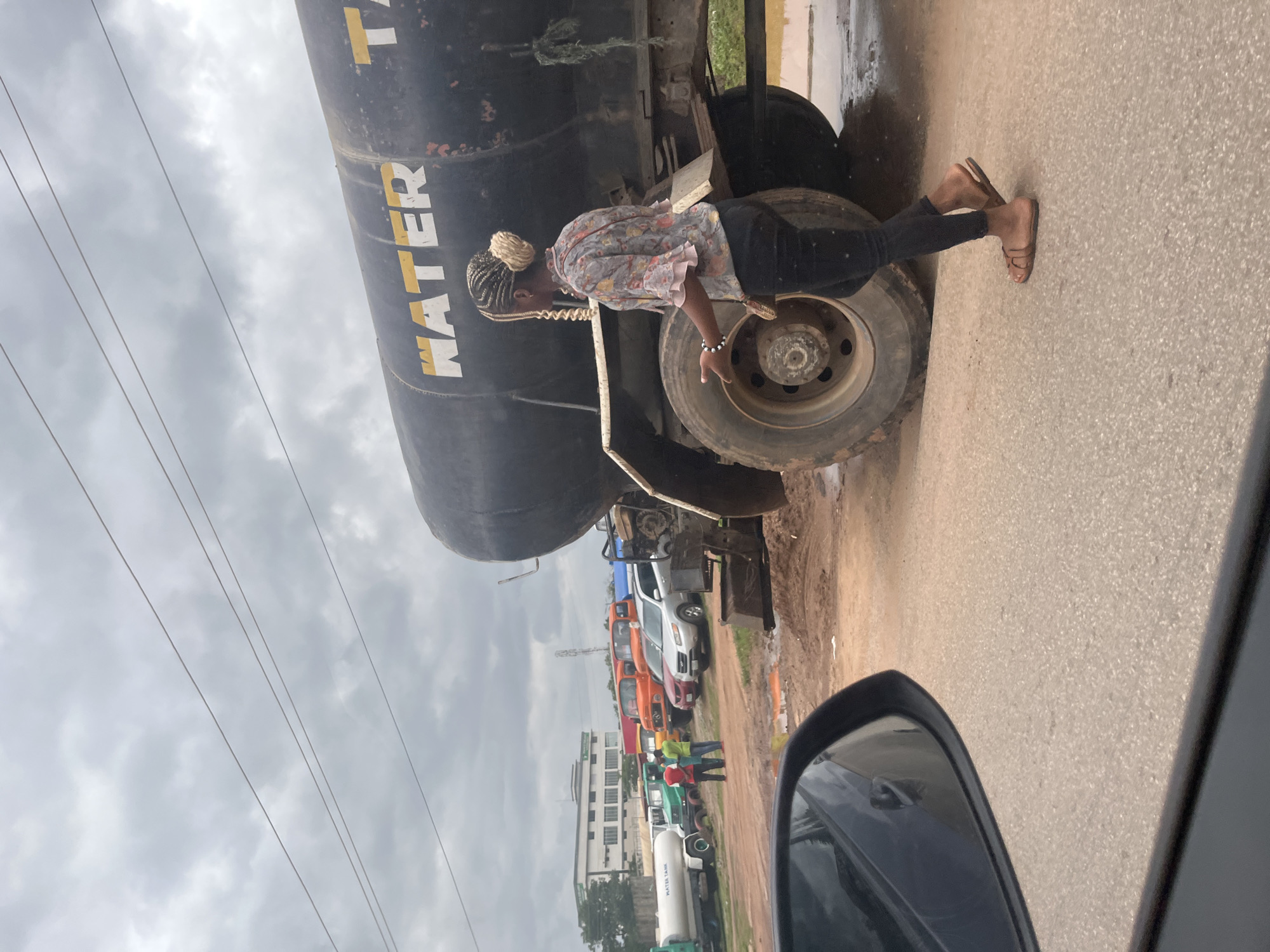 In Galadimawa Village, Nigeria, a young woman with braided hair is walking towards the right, away from a large black water tanker truck. She is wearing a floral print blouse with ruffled sleeves and black pants, and she has on brown sandals. The tanker truck has the word "WATER" painted in yellow and white on its side. Behind the tanker, several other vehicles are parked, including orange trucks and a white water truck labeled "WATER TANK." In the background, there is a modern building and more parked vehicles. The ground is unpaved and appears wet in places, suggesting recent rain. The sky is overcast with clouds. The image is taken from inside a vehicle, with a car's side mirror visible in the foreground on the left.