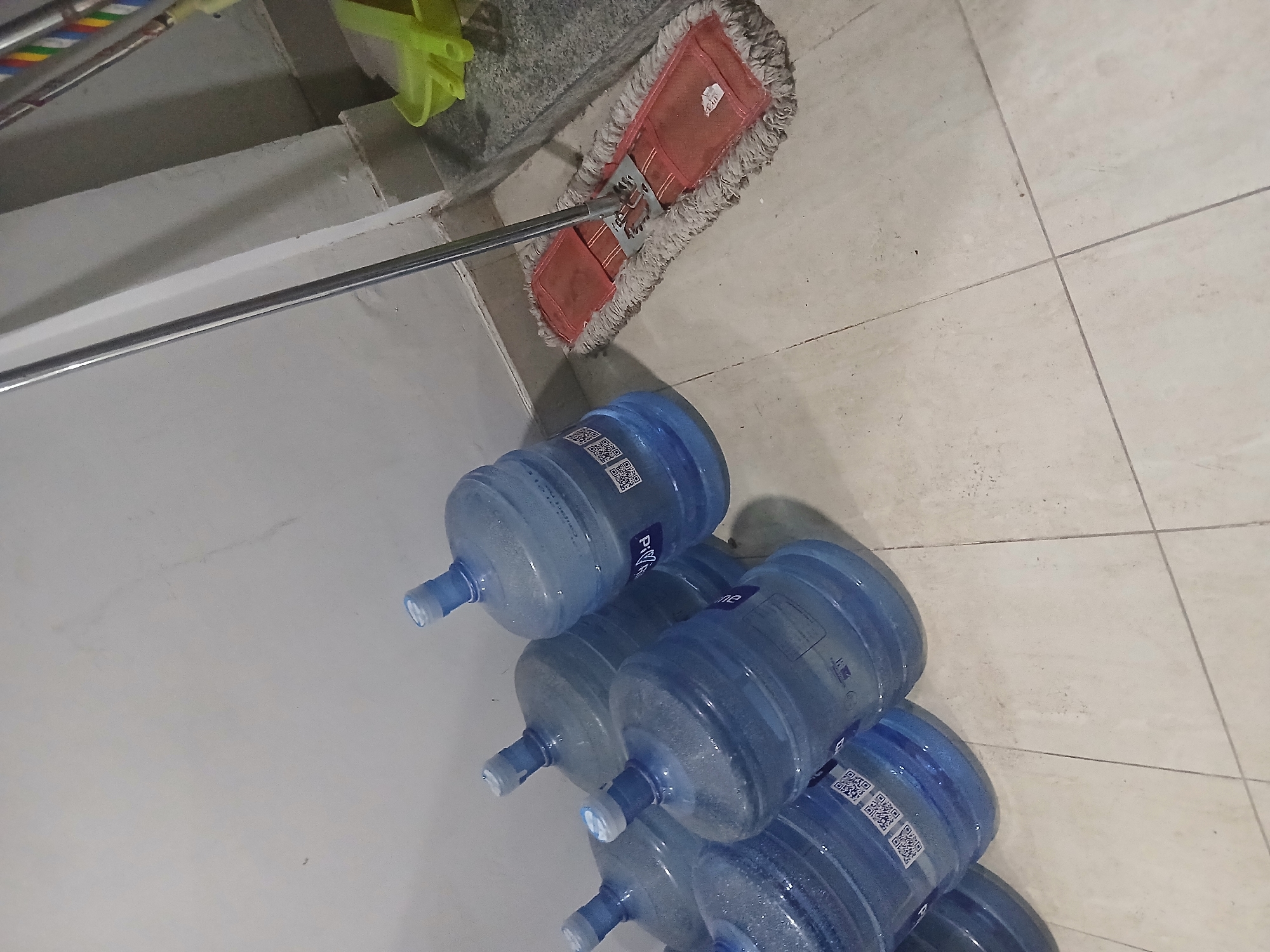 The image shows a group of large, clear plastic water jugs, some partially filled, lined up against a light-colored wall. A mop with a red cleaning pad and a long metal handle is positioned in front of some of the jugs, resting on a light-colored tiled floor. To the right of the mop, a bright green dustpan is visible. The environment appears to be indoors, possibly a storage area or a room where cleaning supplies are kept. The location is not discernible from the image.