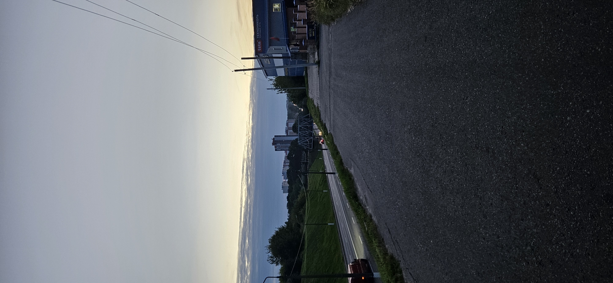 The image depicts an evening scene in Vitsebsk, Belarus, looking east. A paved road curves to the left, with a grassy embankment on its left side. Beyond the embankment, a two-lane road runs parallel, with vehicles visible. In the distance, a railway bridge spans across the road, and beyond that, several multi-story buildings form the city skyline. To the right of the road, a dark-colored, modern building with a signage that reads "ТЕХНОНИКОЛЬ" (Tekhnokol) is situated. Further to the right, there is a smaller brick building with a sign that reads "ЮРИКАС" (Yurikas). Power lines traverse the upper right portion of the image against a pale sky with wispy clouds. The overall impression is one of an urban industrial area at dusk.
