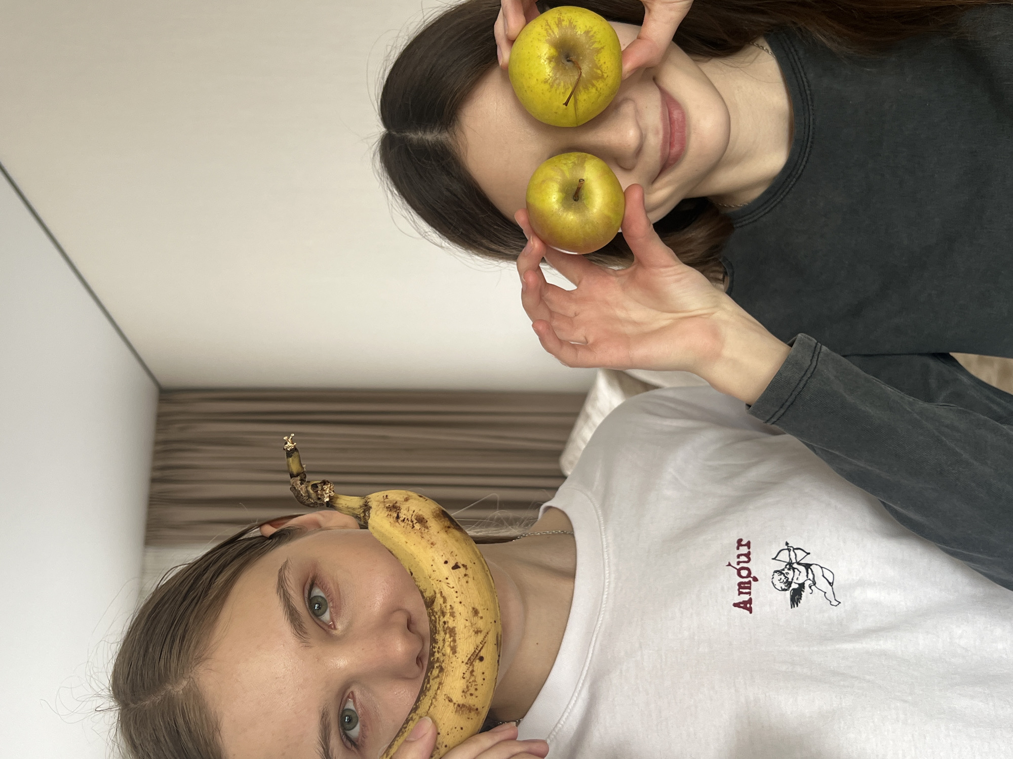 Two women pose playfully. The woman in the upper part of the image holds two yellow apples in front of her eyes. She has dark hair and wears a dark gray long-sleeved shirt. The woman below has a banana held in front of her mouth. She wears a white shirt with a cupid and the word "Amour" printed in burgundy. The background appears to be an interior space with white walls and potentially a bed or other furniture. There is no information to indicate the location is Votkinsk, Russia.