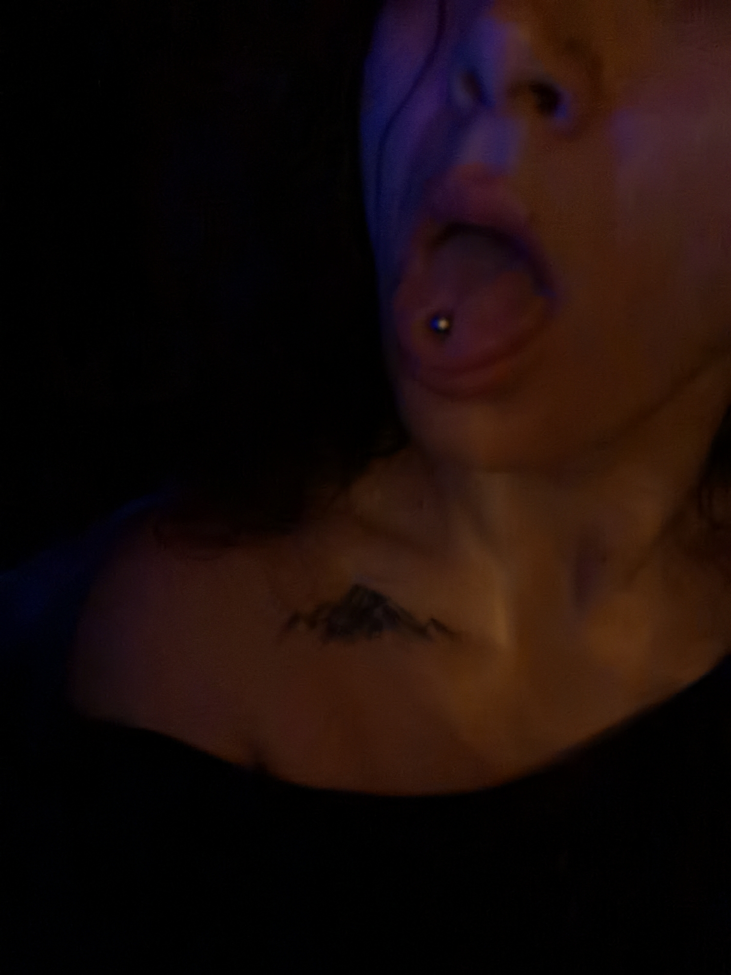 The image shows a person's face, neck, and upper chest. The person has their mouth open, revealing a tongue piercing. A tattoo of a mountain range is visible on their chest. The lighting is dim and has a blue hue. The location of the photo is unknown. The city of Chaikovsky, Russia is not identifiable in the image.
