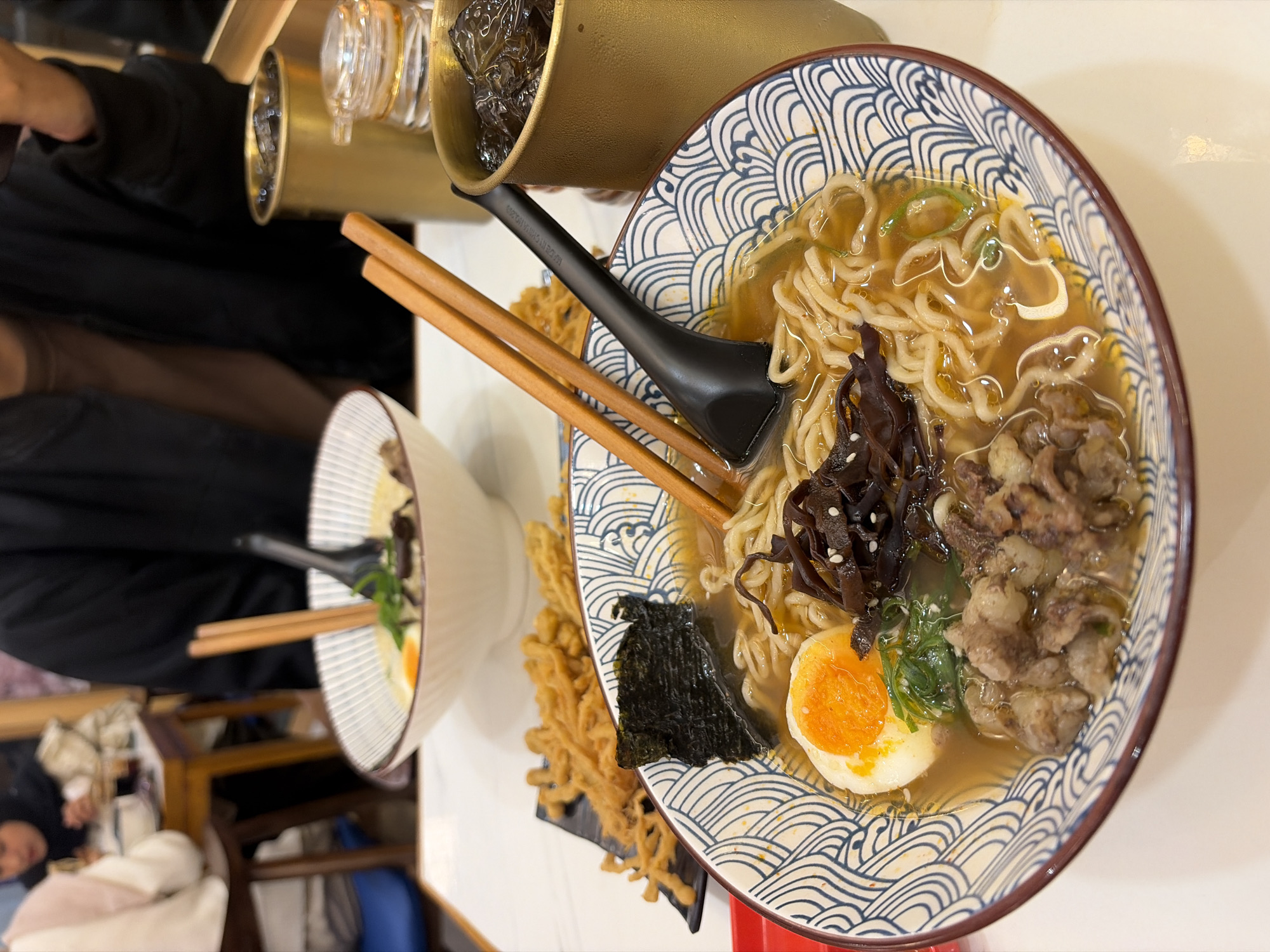 In a restaurant setting, a close-up shot captures two bowls of ramen noodles. The bowl in the foreground is brimming with broth, noodles, thinly sliced beef, a soft-boiled egg with a vibrant orange yolk, seaweed, and dark mushrooms. Chopsticks and a black ladle rest on top of the noodles. A small black plate with deep-fried enoki mushrooms is visible in the background. To the right, a golden metal cup filled with ice and a dark beverage sits next to a glass condiment dispenser. In the blurred background, people are seated at tables, suggesting a public dining space. The overall impression is a detailed view of a meal being served in a restaurant. This scene could be located in Pekalongan, Indonesia, a city known for its culinary scene and diverse eateries.